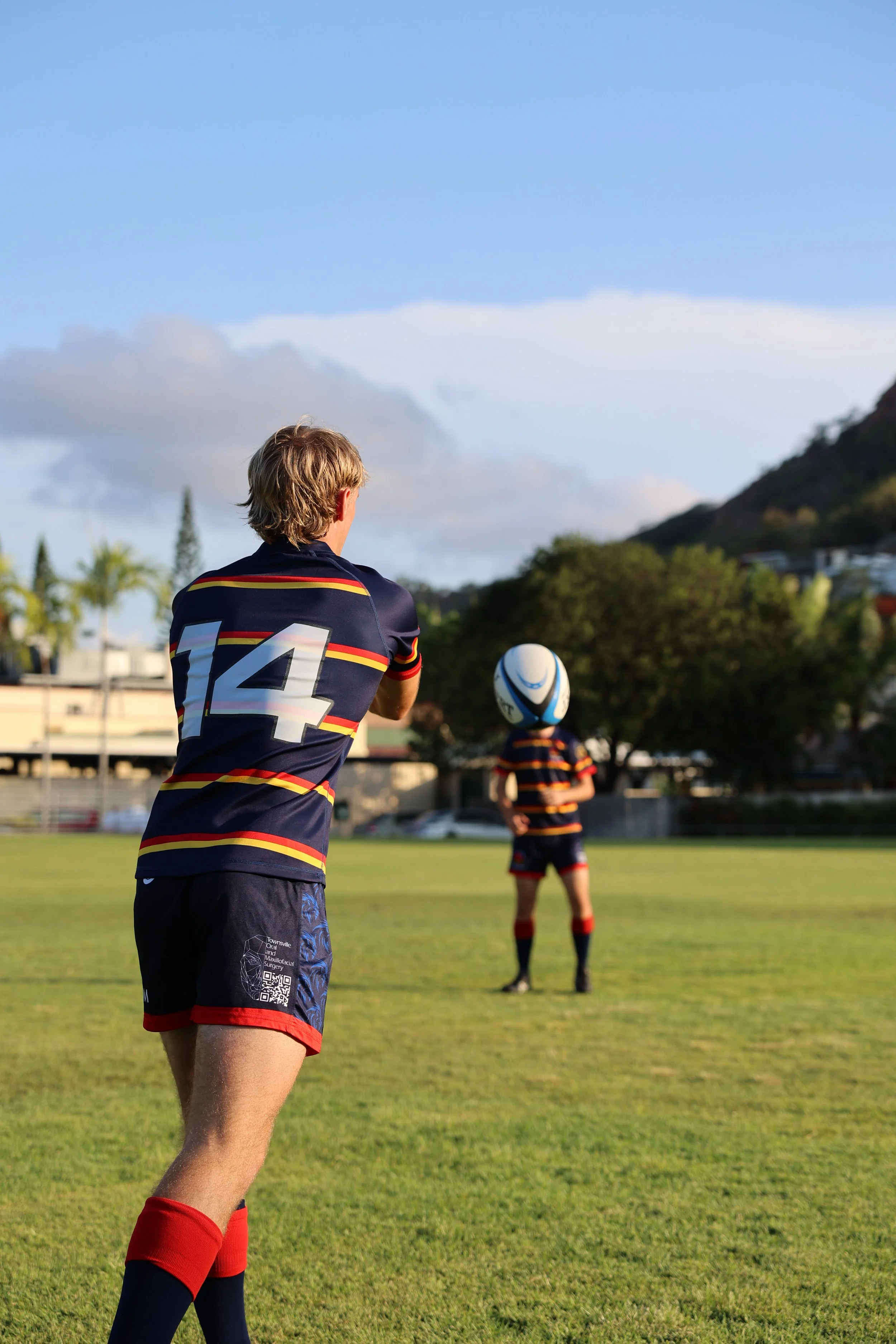 Two young boys in rugby uniforms on a grassy field, one preparing to catch or throw a rugby ball, with trees, houses, and a mountain in the background under a partly cloudy sky.