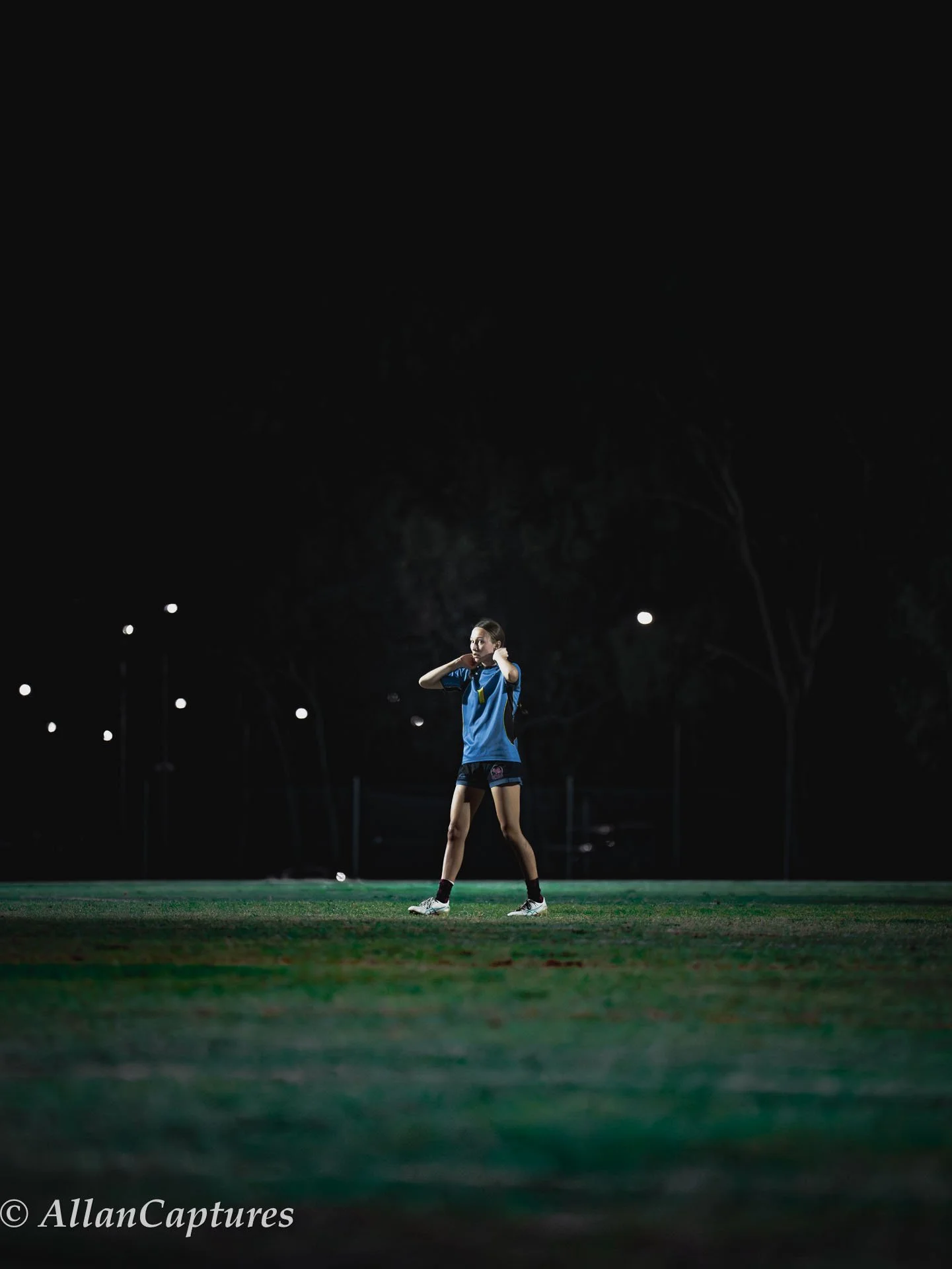 A female athlete standing on a grassy sports field at night, wearing a blue sports uniform, adjusting her headset, with bright field lights in the background.