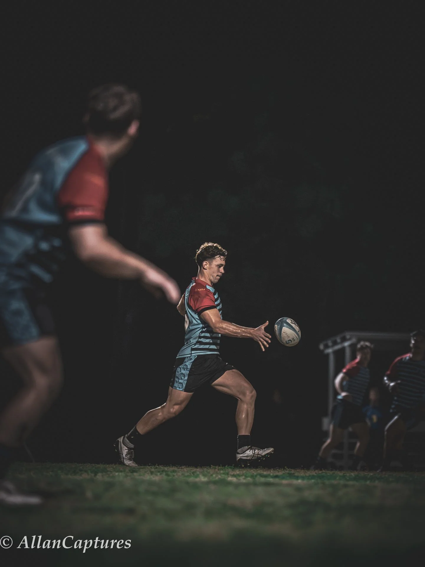 Rugby players warm-up on the field at night, focusing on a player passing a rugby ball.