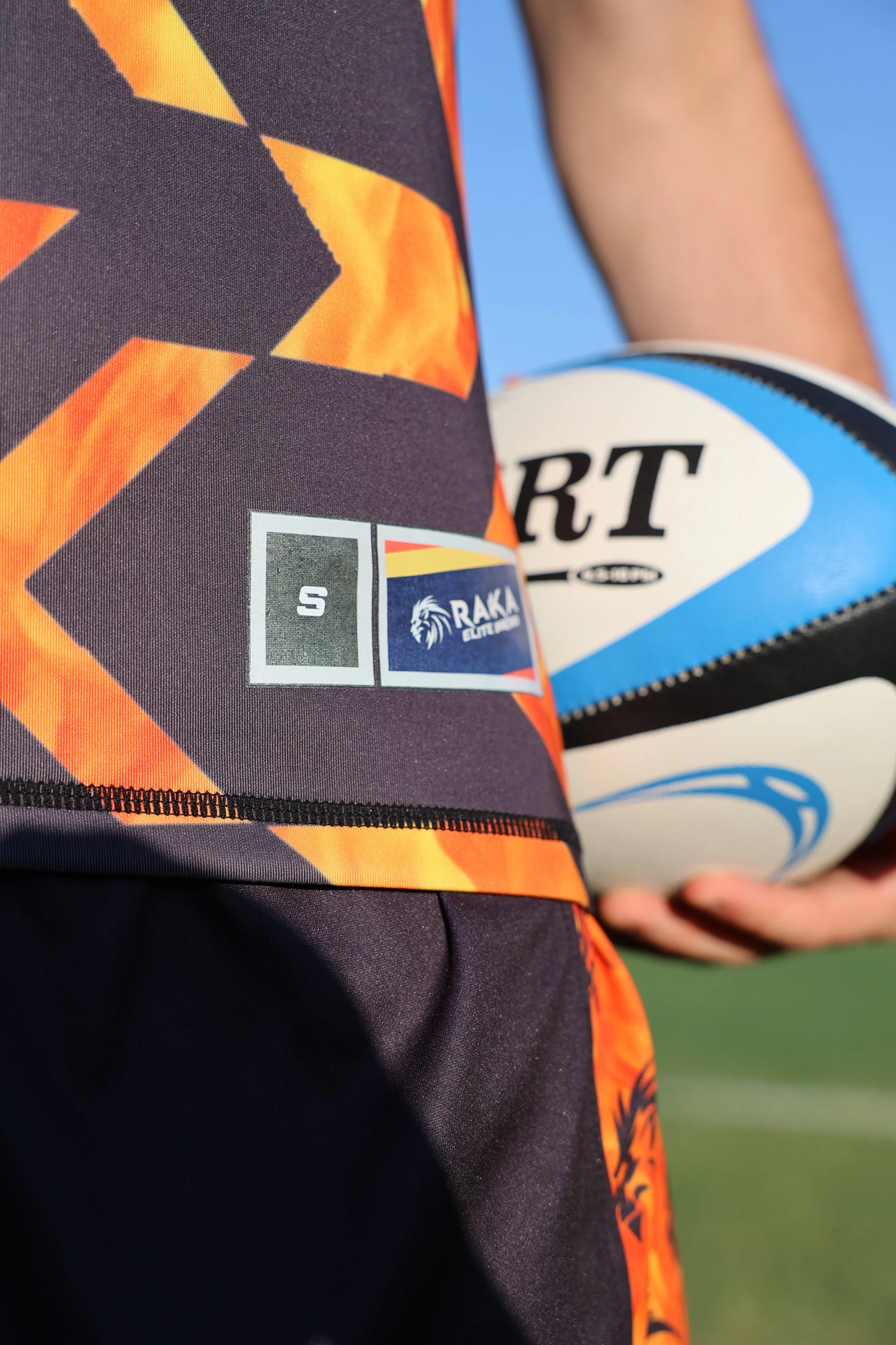 Close-up of a person holding a rugby ball in front of their shorts, which feature a logo and color pattern. The background is clear blue sky.
