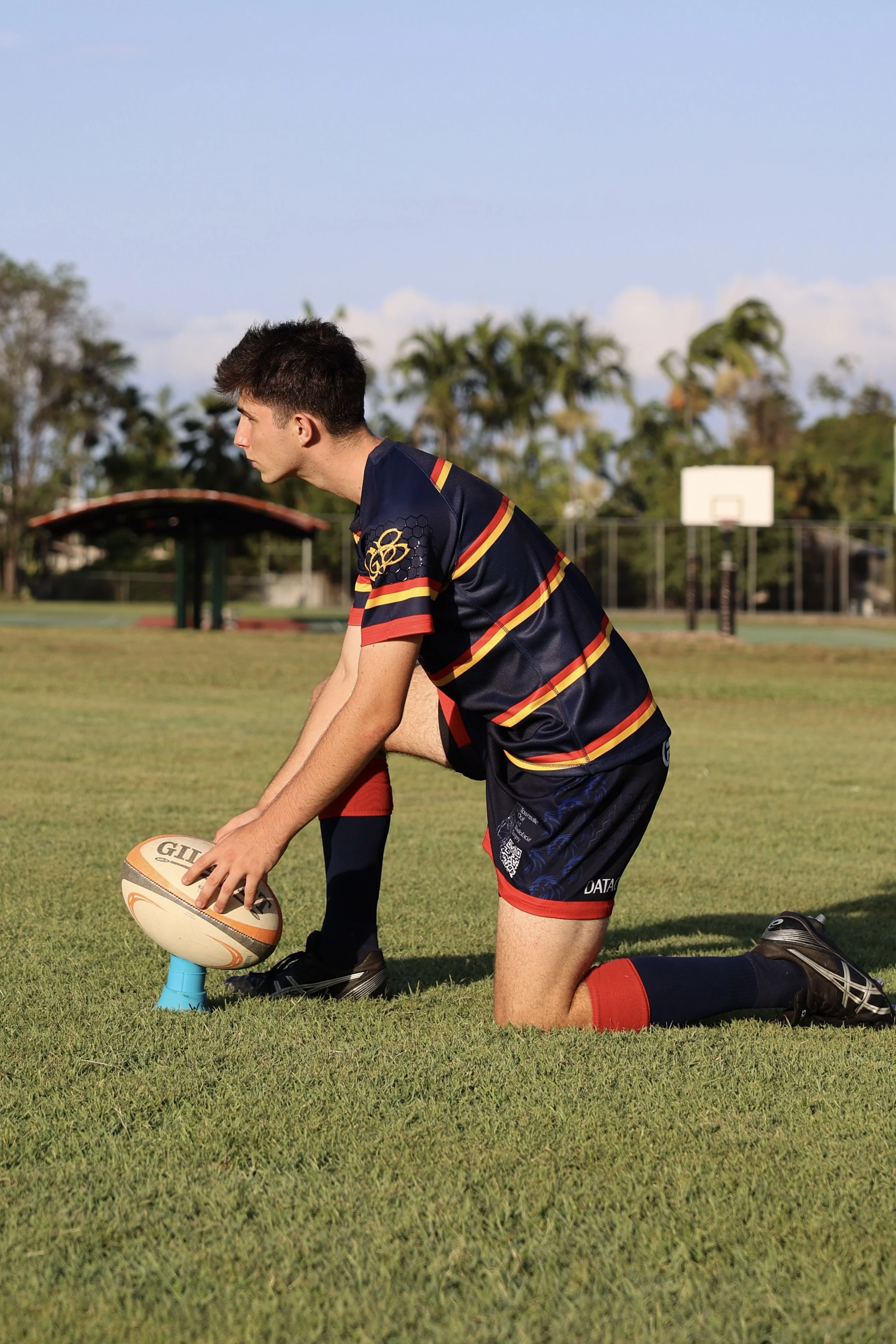A young male rugby player is kneeling on a grassy field, preparing to place a rugby ball on a kicking tee. He is wearing a navy blue and red rugby uniform, with a view of a sports court and trees in the background.
