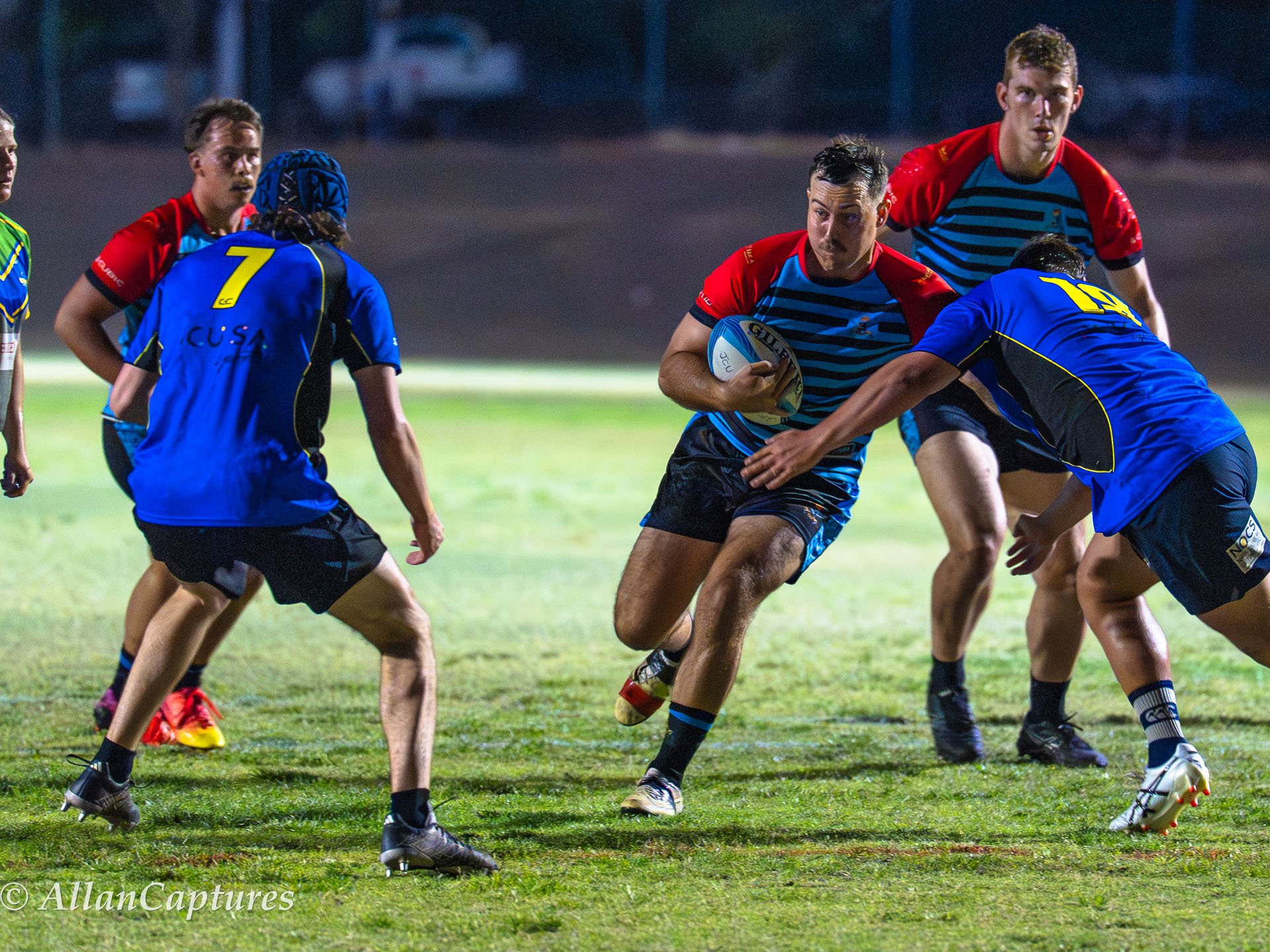 Rugby players in action on the field during a night game. One player in a striped blue and black jersey is running with the ball, while others in blue and red jerseys are positioned to block or tackle.