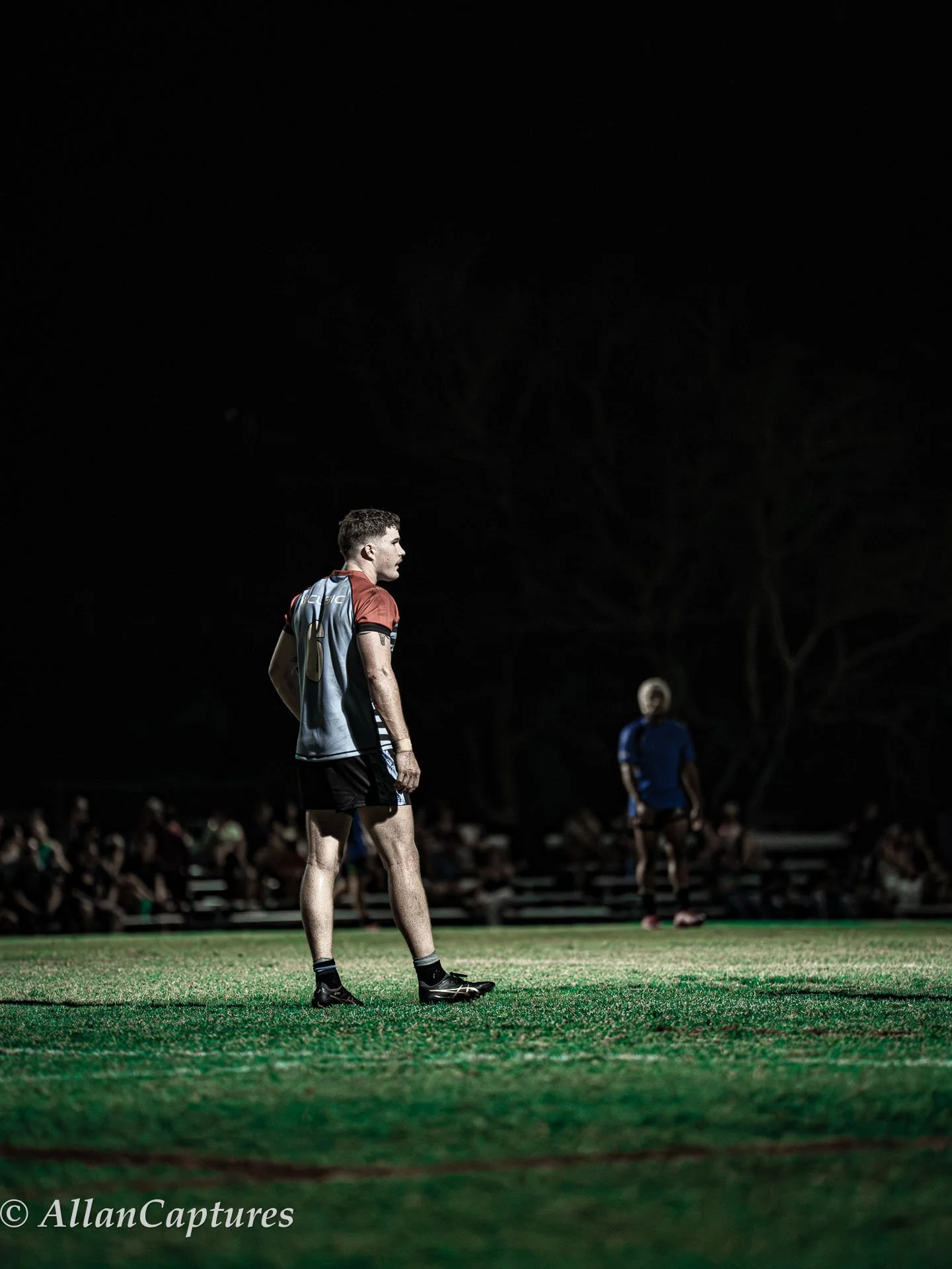 A soccer player stands on the field at night, illuminated by stadium lights, with a dark background and spectators in the distance.