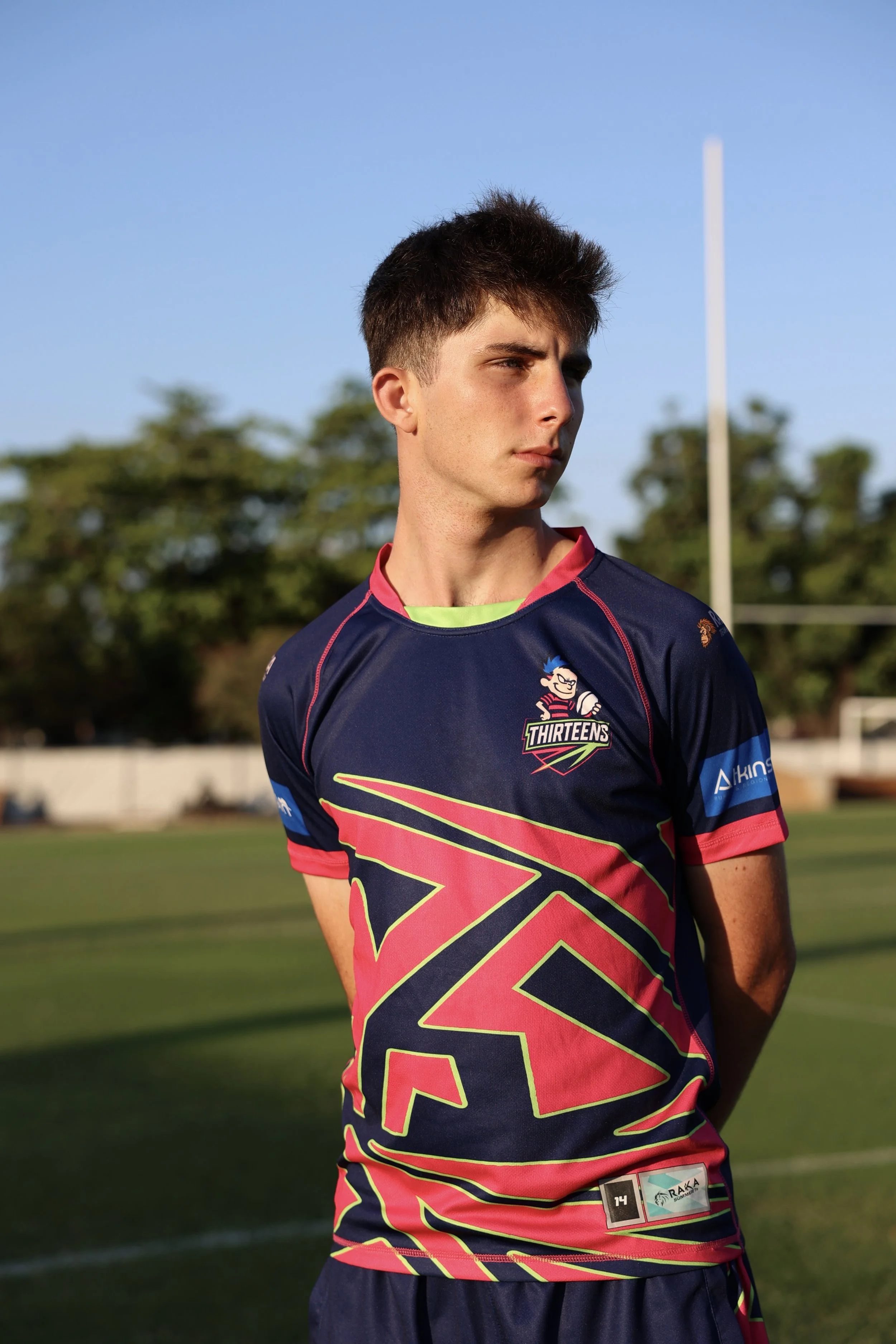 A young male rugby player standing on a field, wearing a colorful jersey with 'Thirteens' logo, under a clear blue sky with trees in the background.