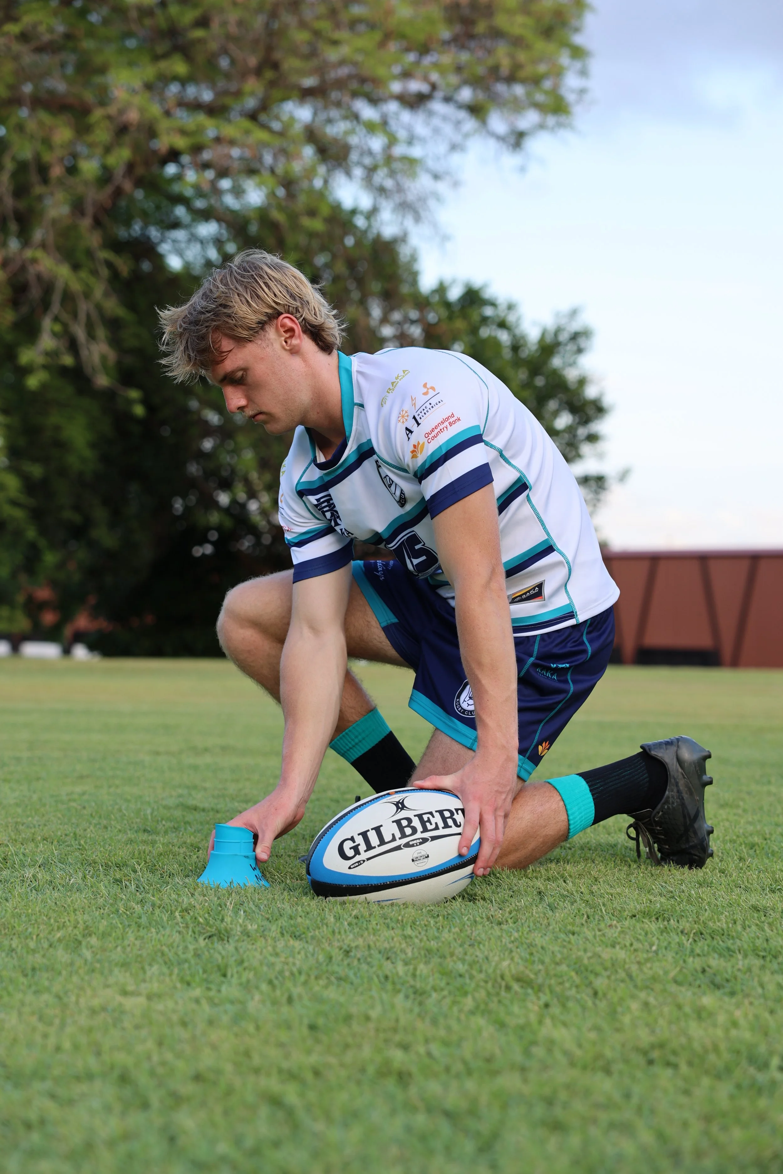 A young man in a rugby uniform kneels on a grassy field, placing a rugby ball and a blue cone in front of him for practice.