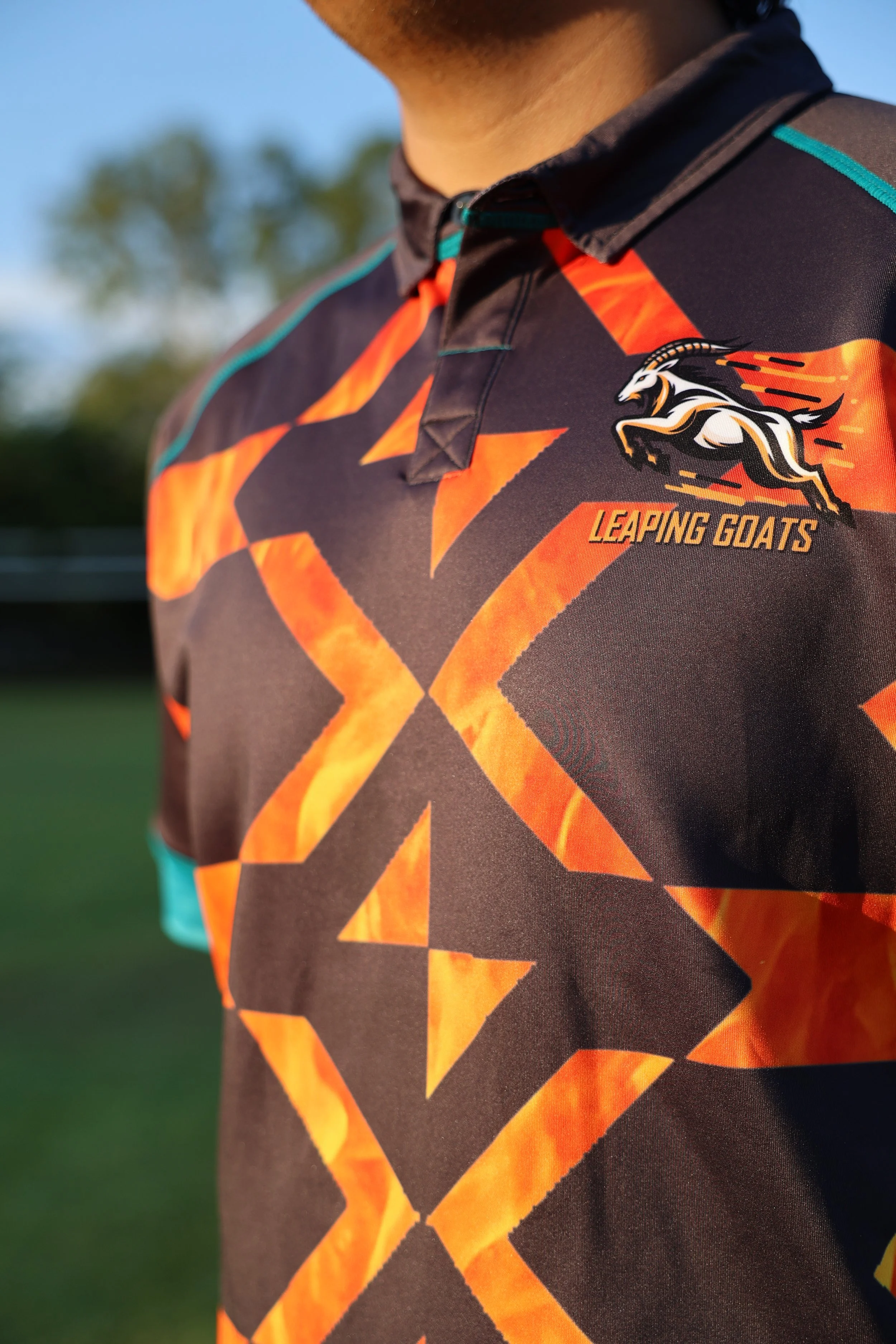 Close-up of a sports jersey featuring a leaping goat mascot and the text "Leaping Goats" on the upper right side. The jersey has a black base with orange flame-like geometric patterns and teal accents.