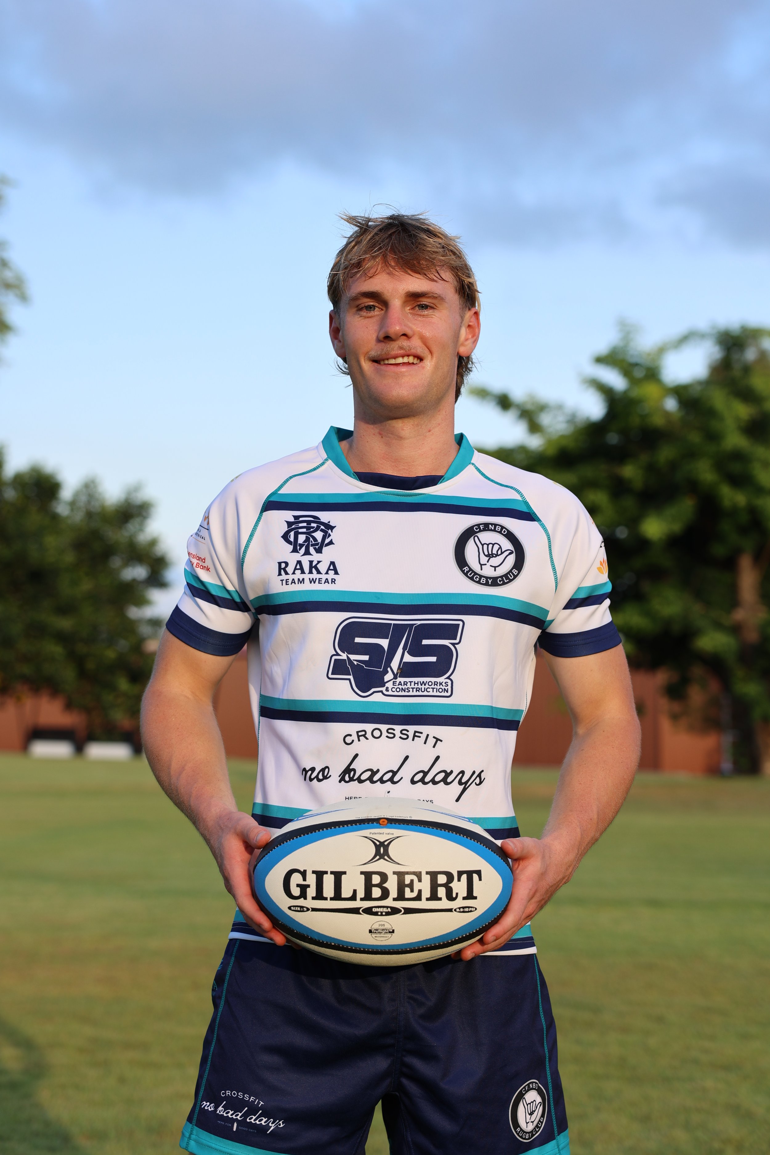 A young man in a rugby jersey holding a Gilbert rugby ball outdoors on a grassy field, with trees and a partly cloudy sky in the background.