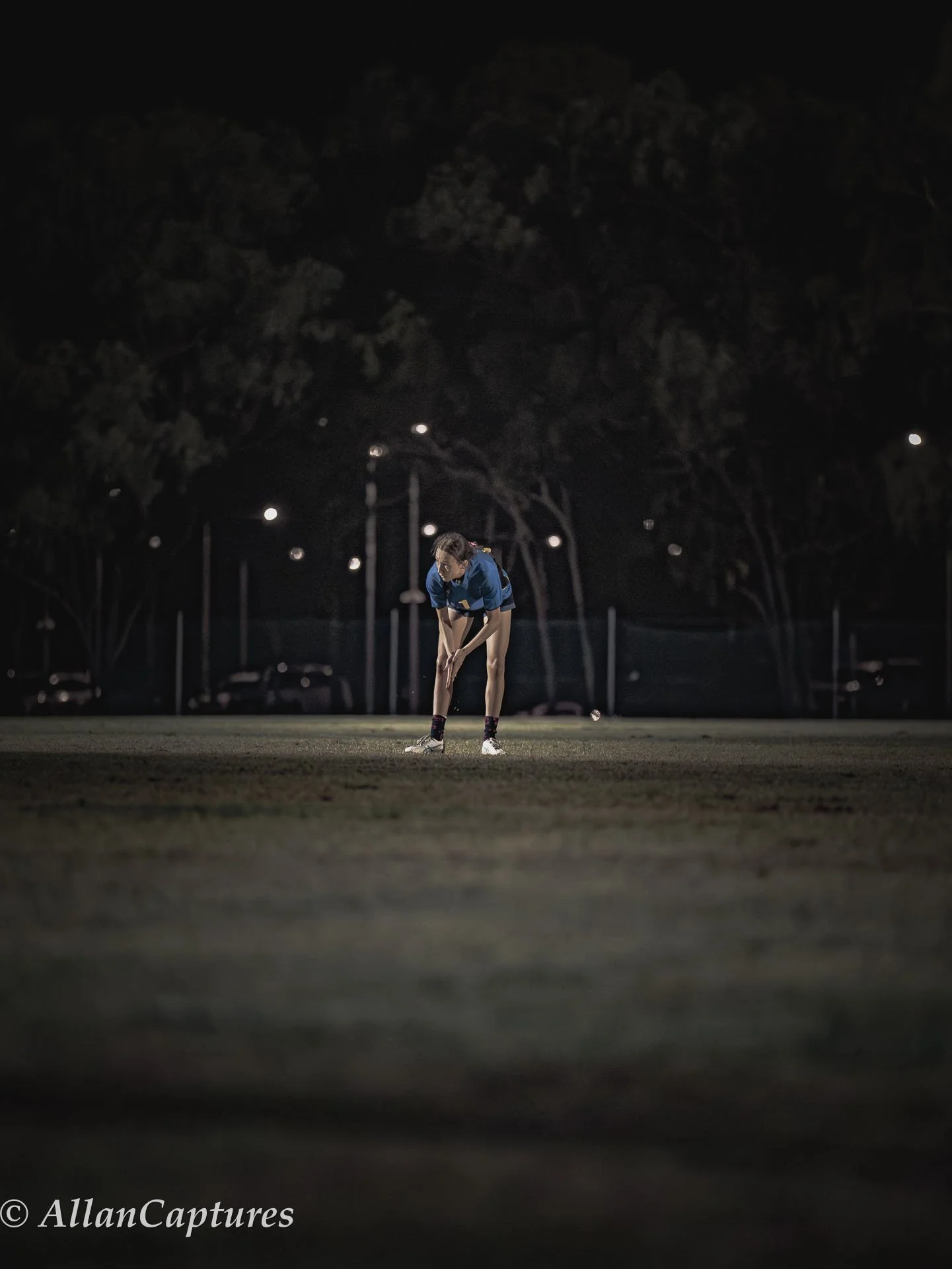 A female athlete is preparing to hit a golf ball at night on a sports field under bright lights, with trees and parked cars in the background.