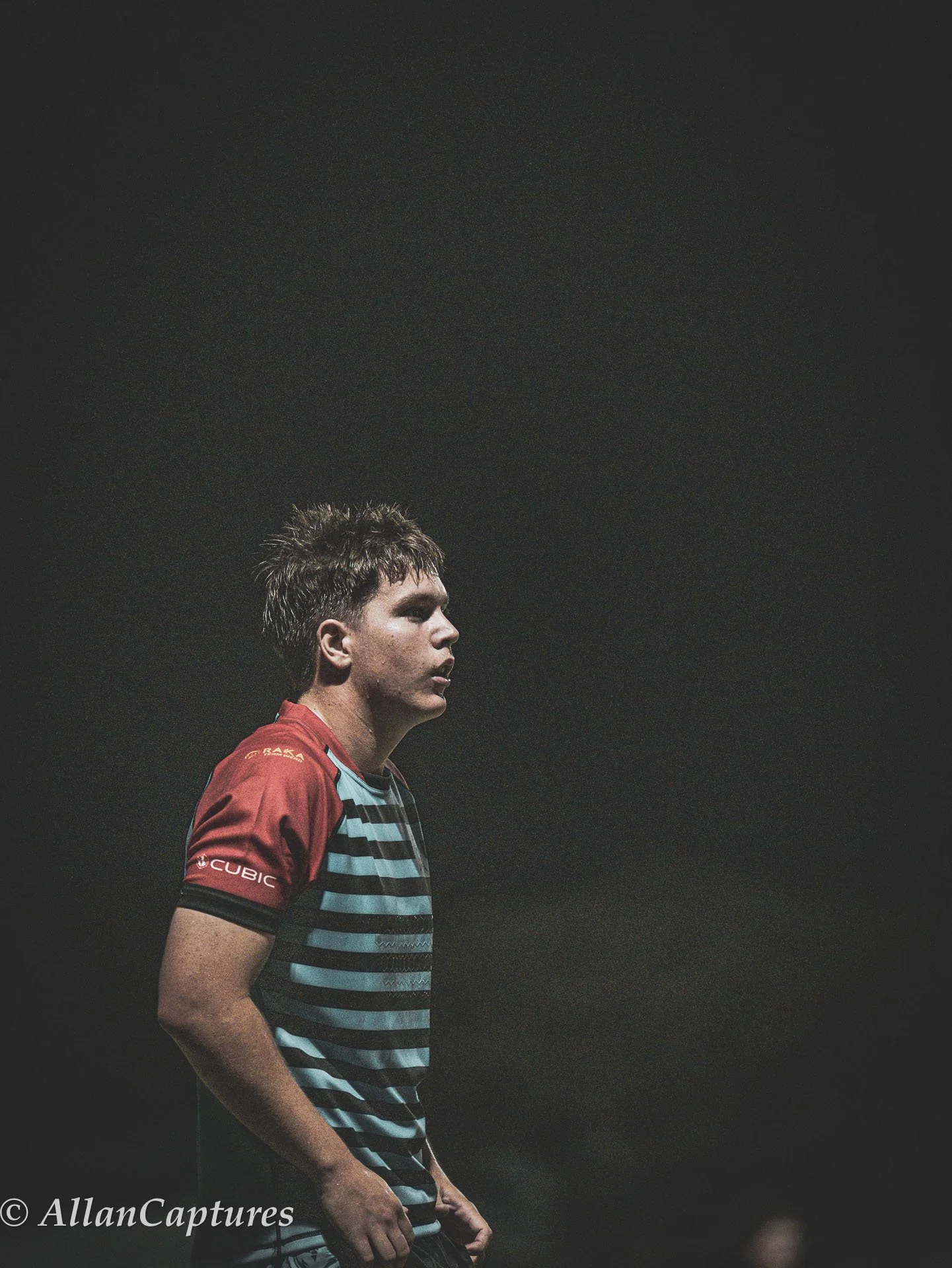 A young man in a striped sports shirt standing outdoors at night with a dark sky behind him.