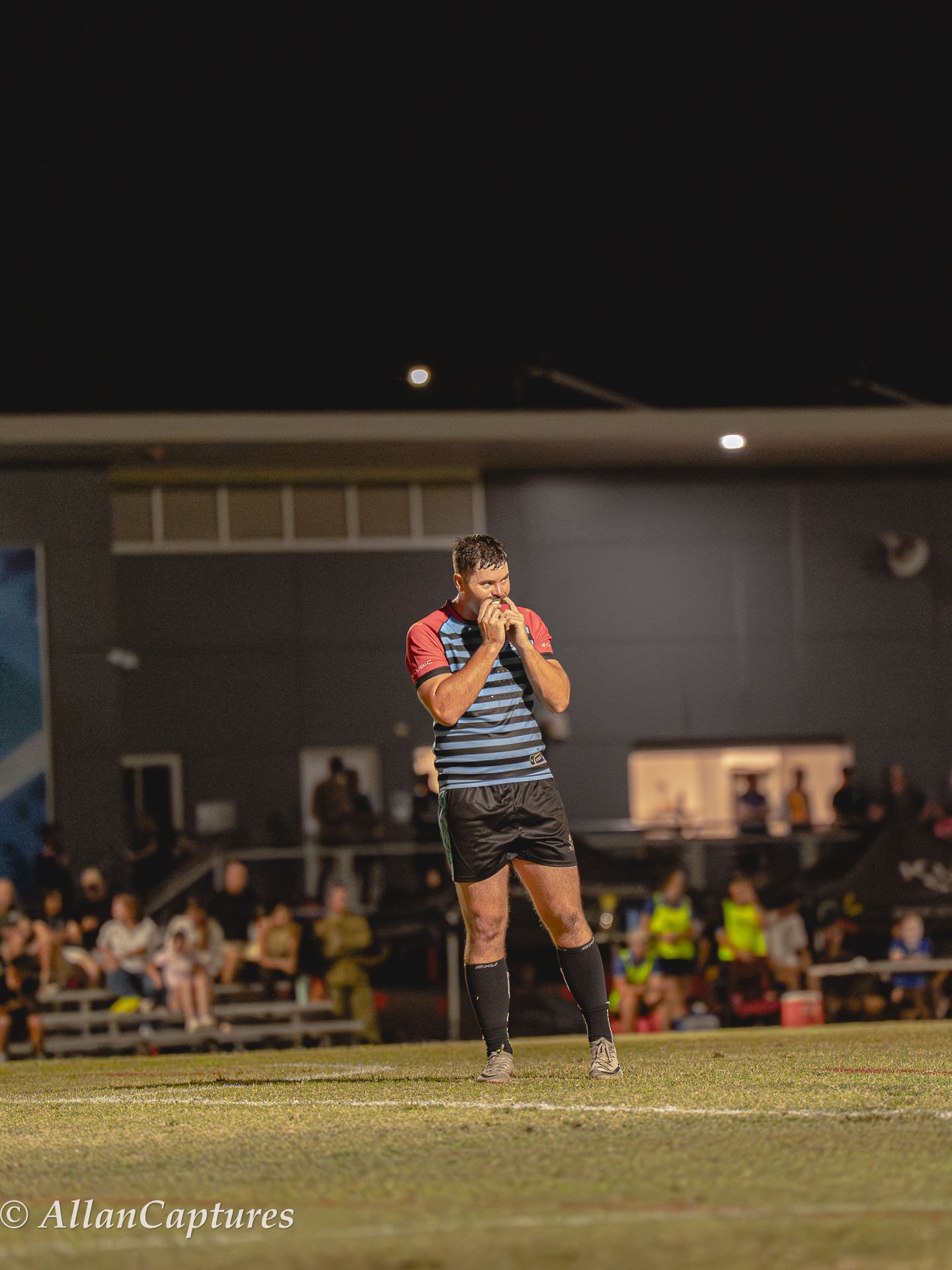 A rugby player in a striped shirt and black shorts standing on the field at night, biting his jersey, with spectators in the background.