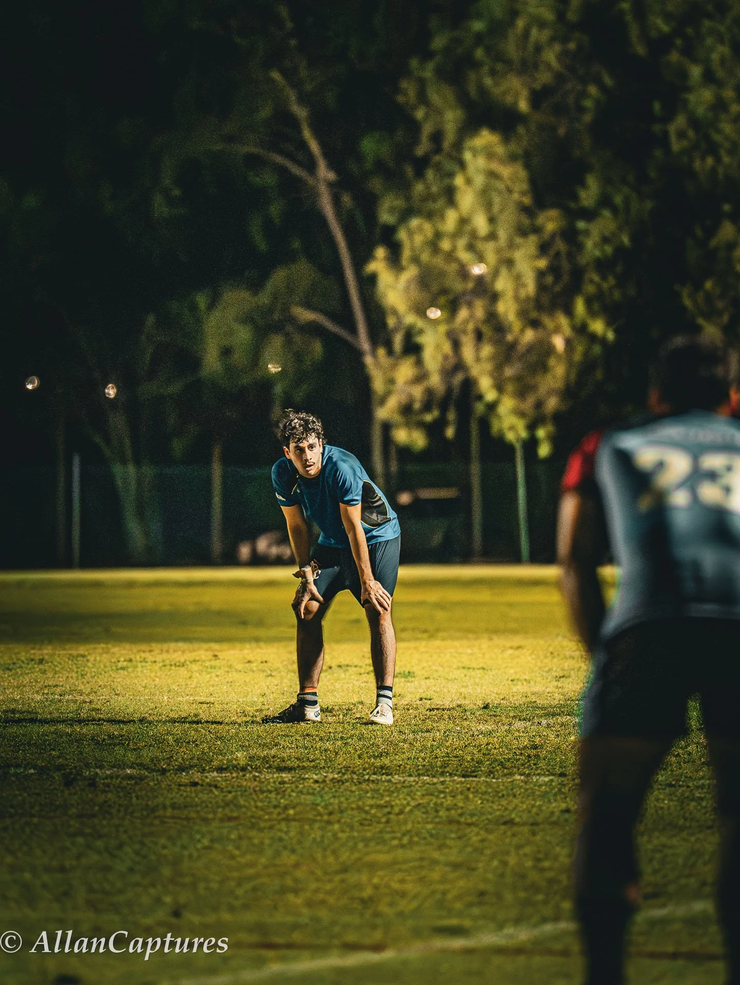 A young man standing on a football field at night, appearing to be exhausted or catching his breath, with another player partially visible in the foreground and trees illuminated in the background.