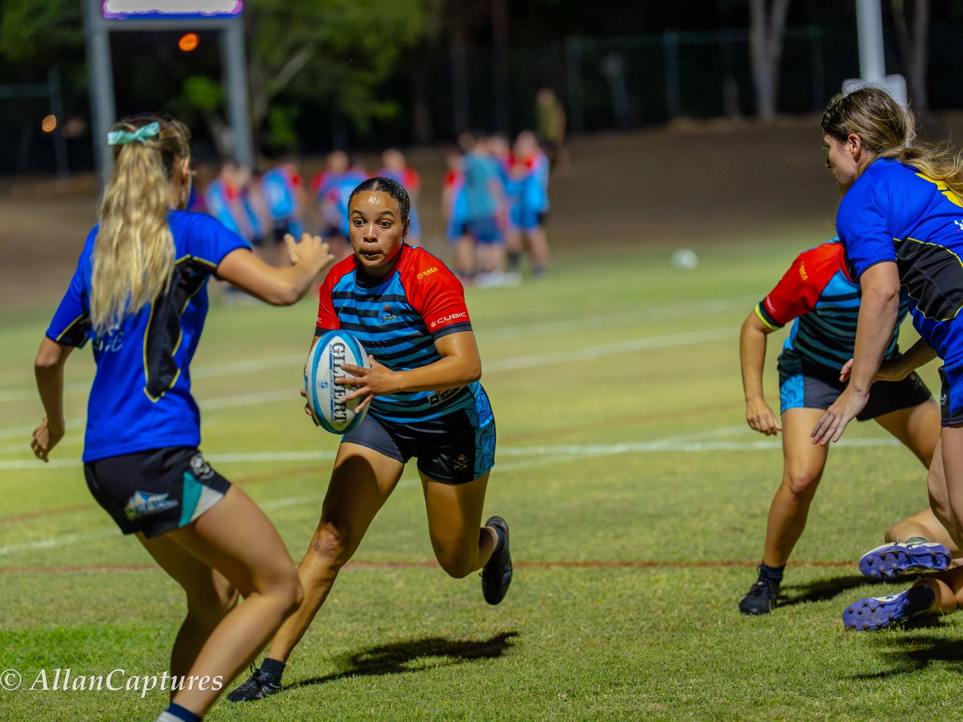 Girls playing rugby on a grassy field at night, with one girl running with the ball while others defend.