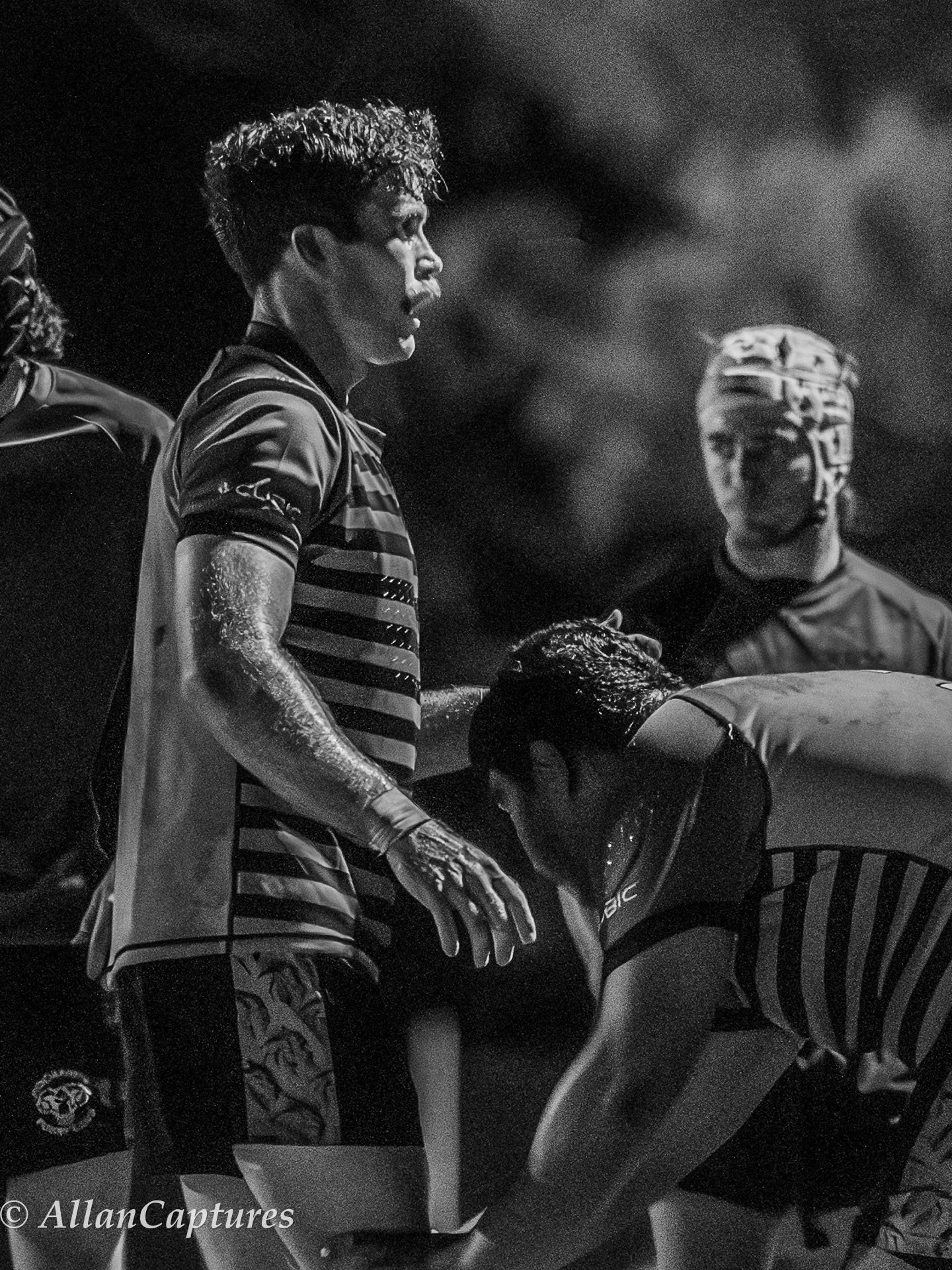 A black and white photo of a rugby player wearing a striped jersey, surrounded by teammates, with one teammate bending over and the others standing nearby.
