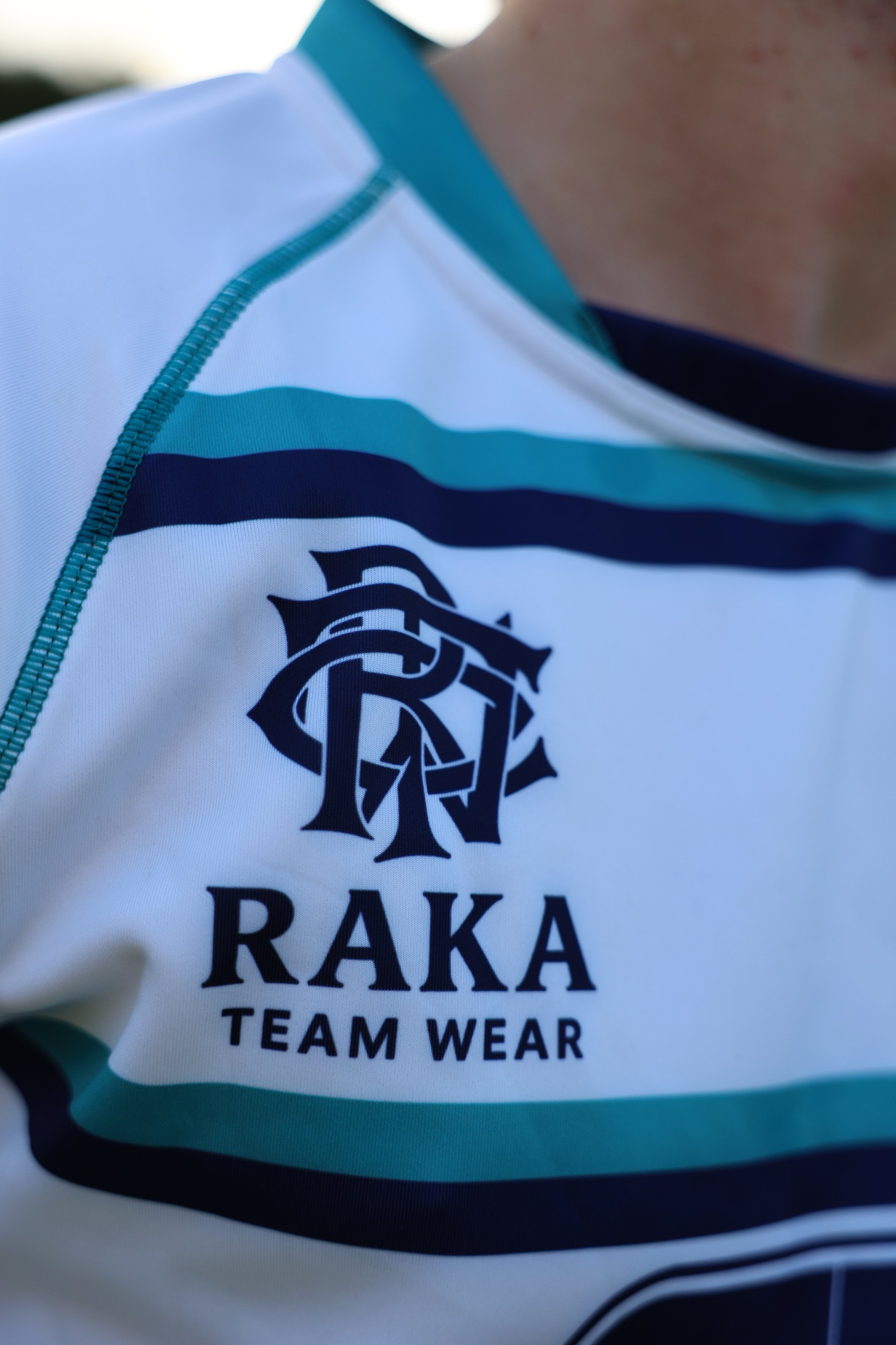 Close-up of a sports jersey with the logo and text 'RAKA TEAM WEAR' on the chest, featuring blue, green, and white colors.