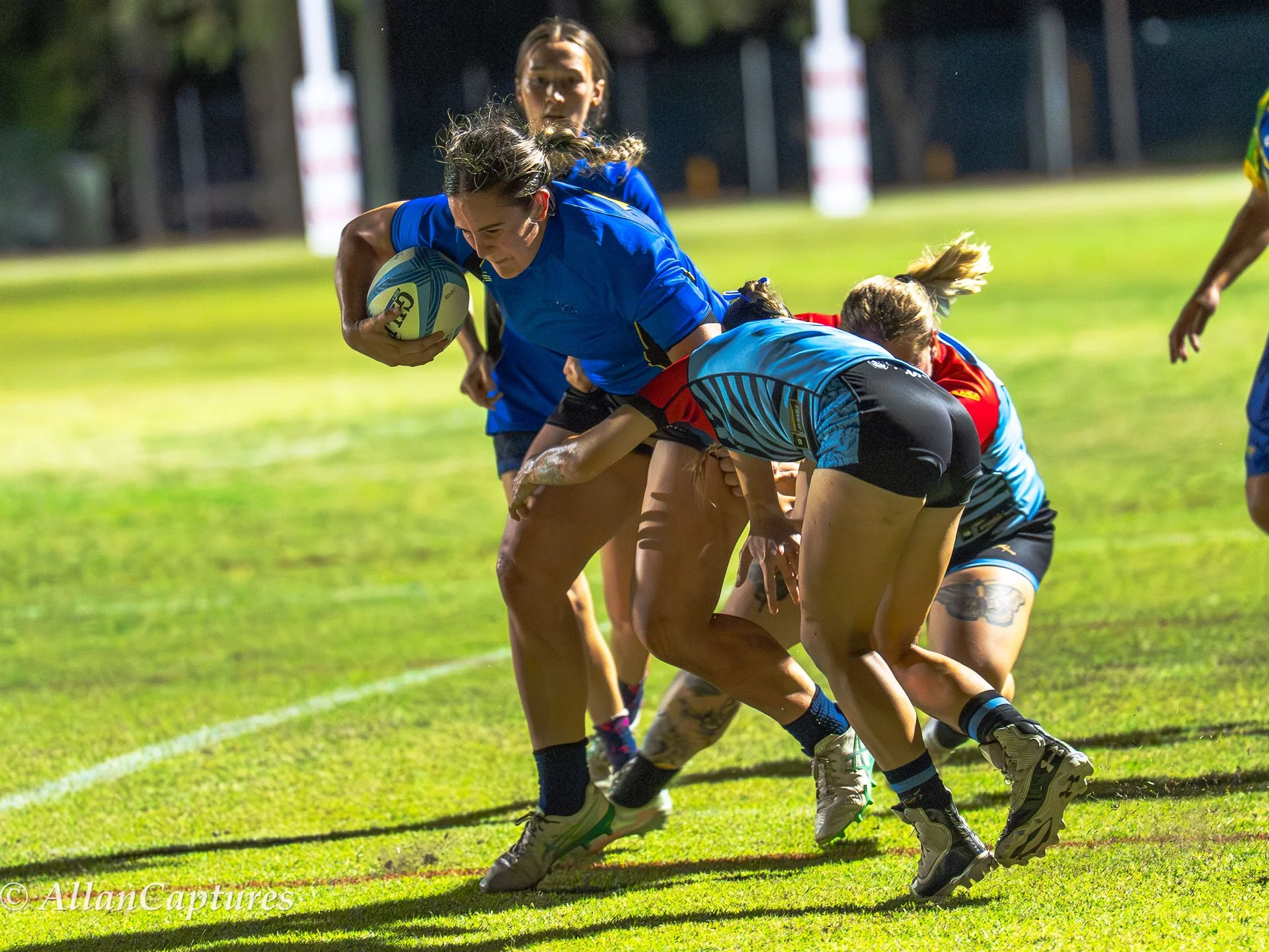 Women playing rugby on a grassy field, with one woman holding a rugby ball and others attempting to tackle her.