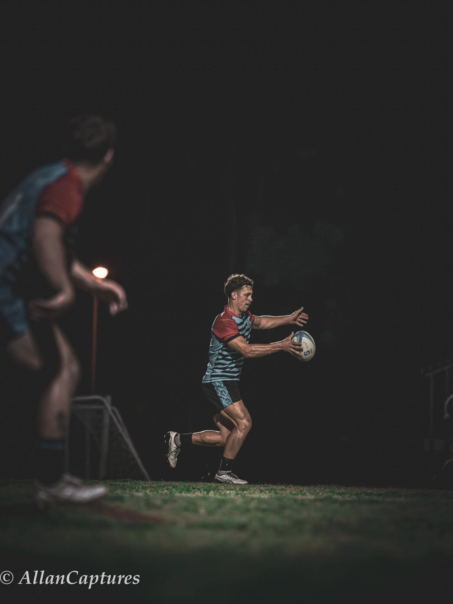 A rugby player holding a ball on a dark field at night, with a teammate partially visible in the foreground.