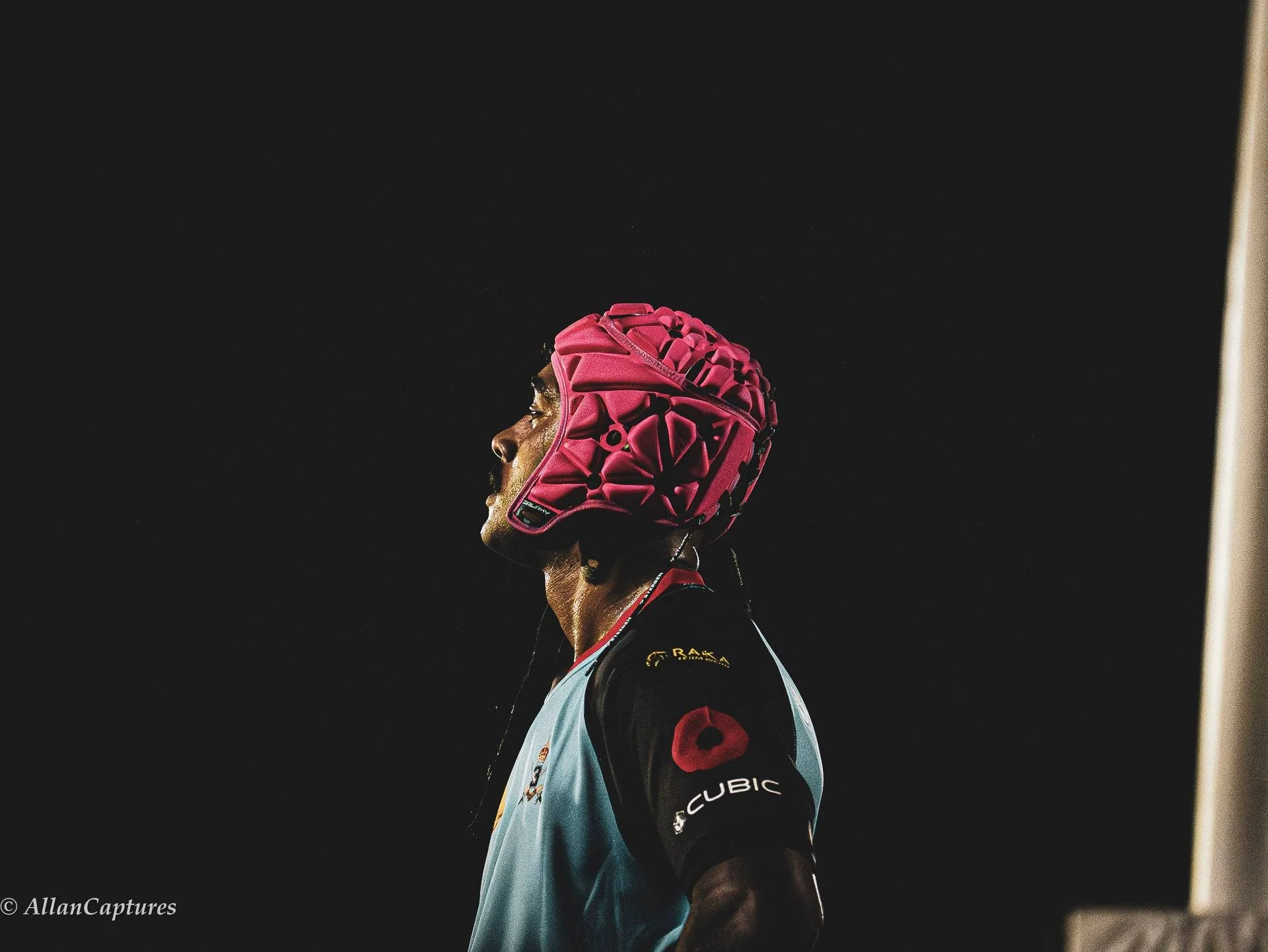 A rugby player wearing a pink helmet and a light blue and black jersey, standing against a dark background, looking to the left.