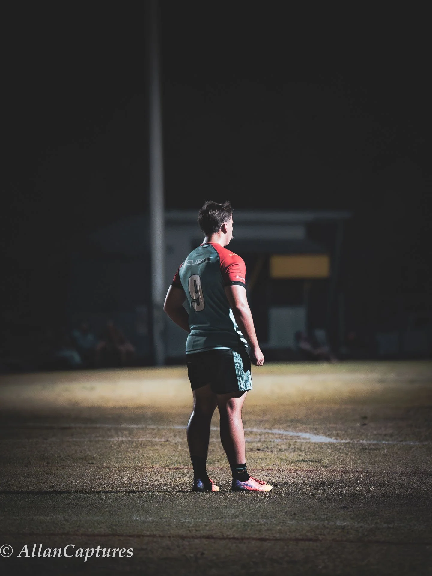 A soccer player wearing a jersey with the number 9 stands alone on a dimly lit outdoor soccer field at night, looking away from the camera.