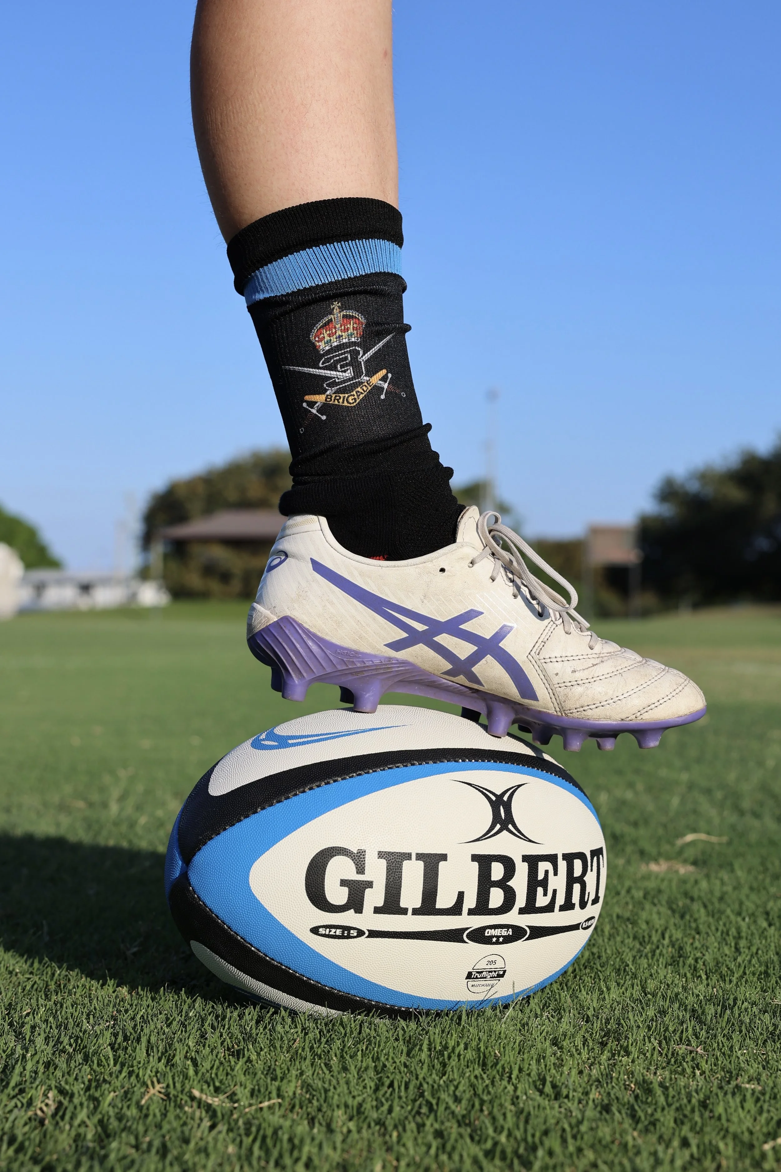 Close-up of a rugby player balancing on a rugby ball on a grassy field, wearing a white and purple cleat, black socks with a logo, and a blue sky background.