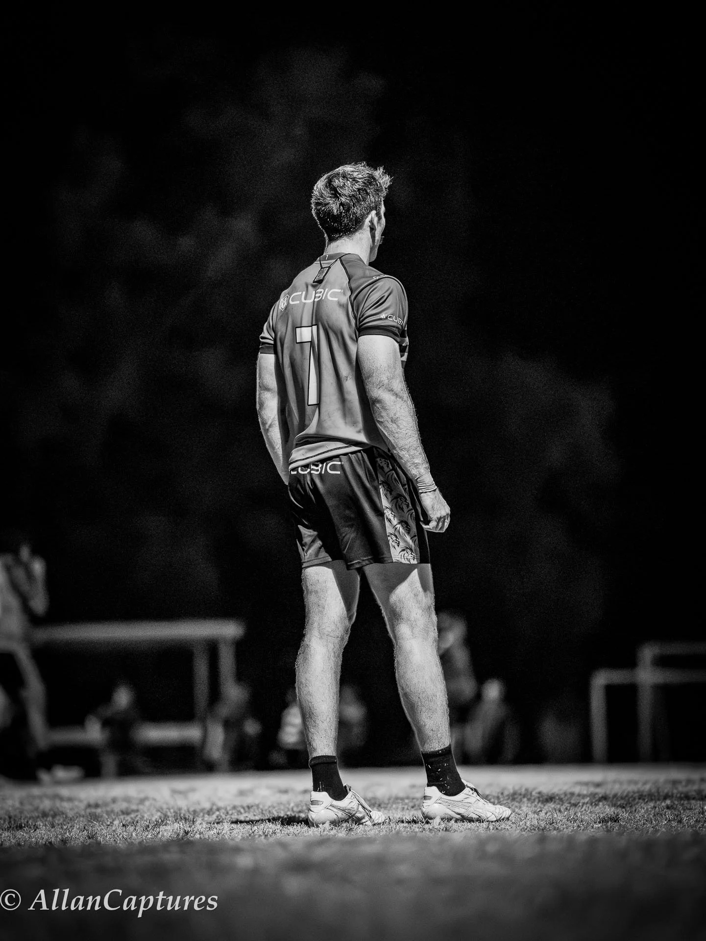 A man in athletic gear stands on a sports field at night, viewed from behind, with blurred figures and equipment in the background.