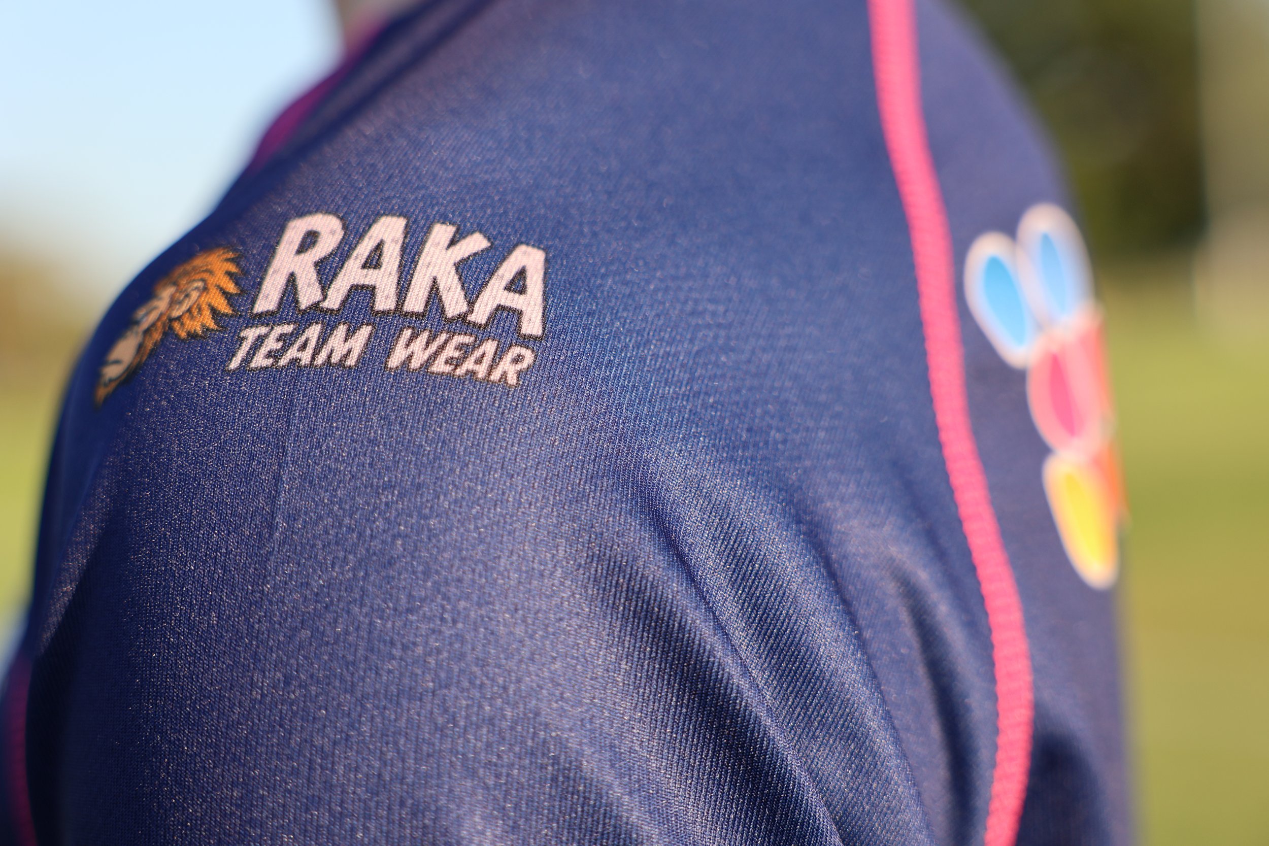 Close-up of a navy blue rugby jersey with pink piping, featuring the RAKA TEAM WEAR logo and a floral emblem, with additional colorful patches on the sleeve.