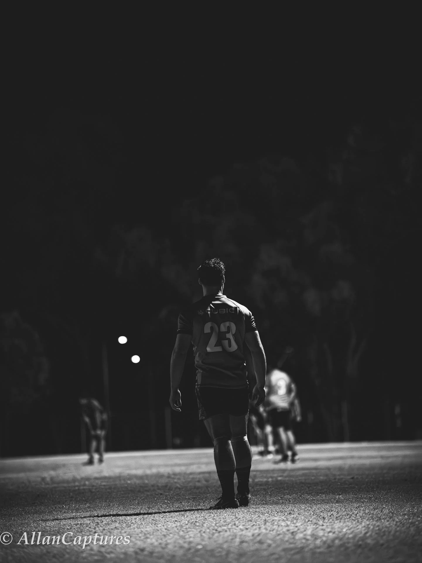 A black-and-white photo of a football player wearing a jersey with the number 23, walking on a field at night. There are other players in the background and trees lining the field.