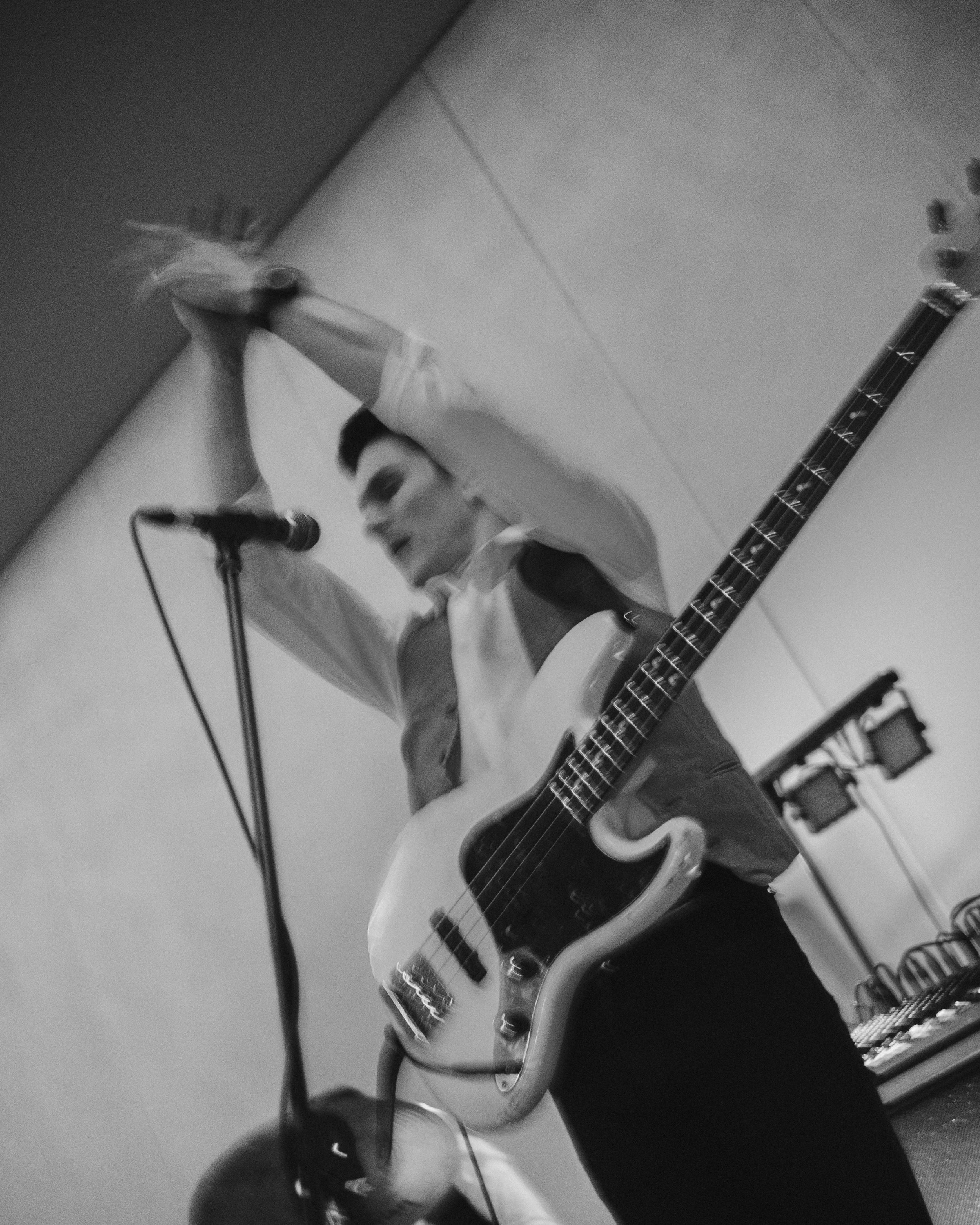 Musician on stage playing bass guitar, clapping hands near microphone in black and white image.