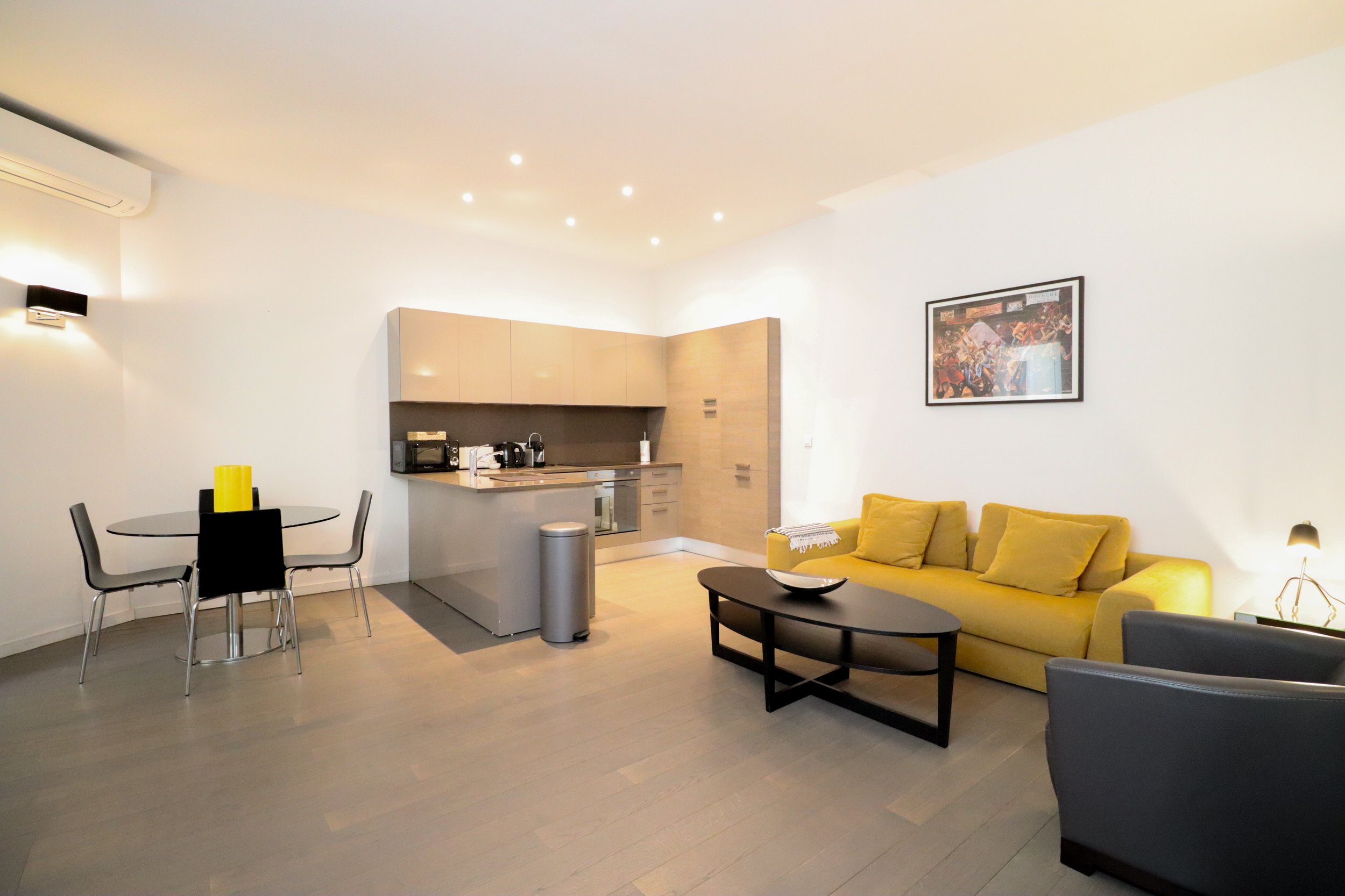 Salon intérieur moderne avec canapé jaune, table basse noire, cuisine avec électroménagers, tableau artistique au mur et éclairage intégré au plafond