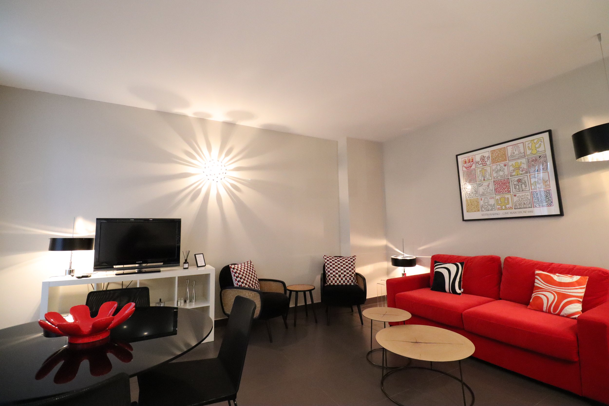 Salon moderne avec un canapé rouge, des tables basses rondes en bois, une télévision, des lampes de table, des chaises noires et décorations murales colorées.