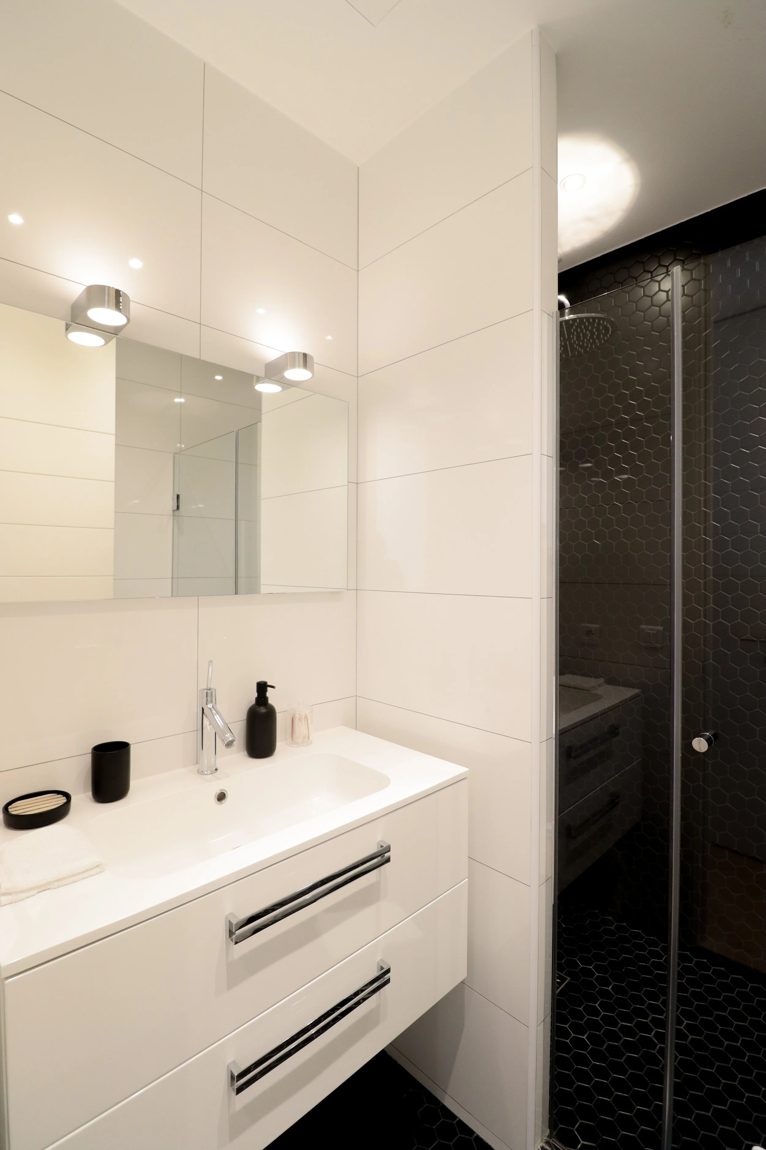 Une salle de bain moderne avec un lavabo blanc, un miroir, et un espace douche avec murs en carreaux noir hexagonaux.