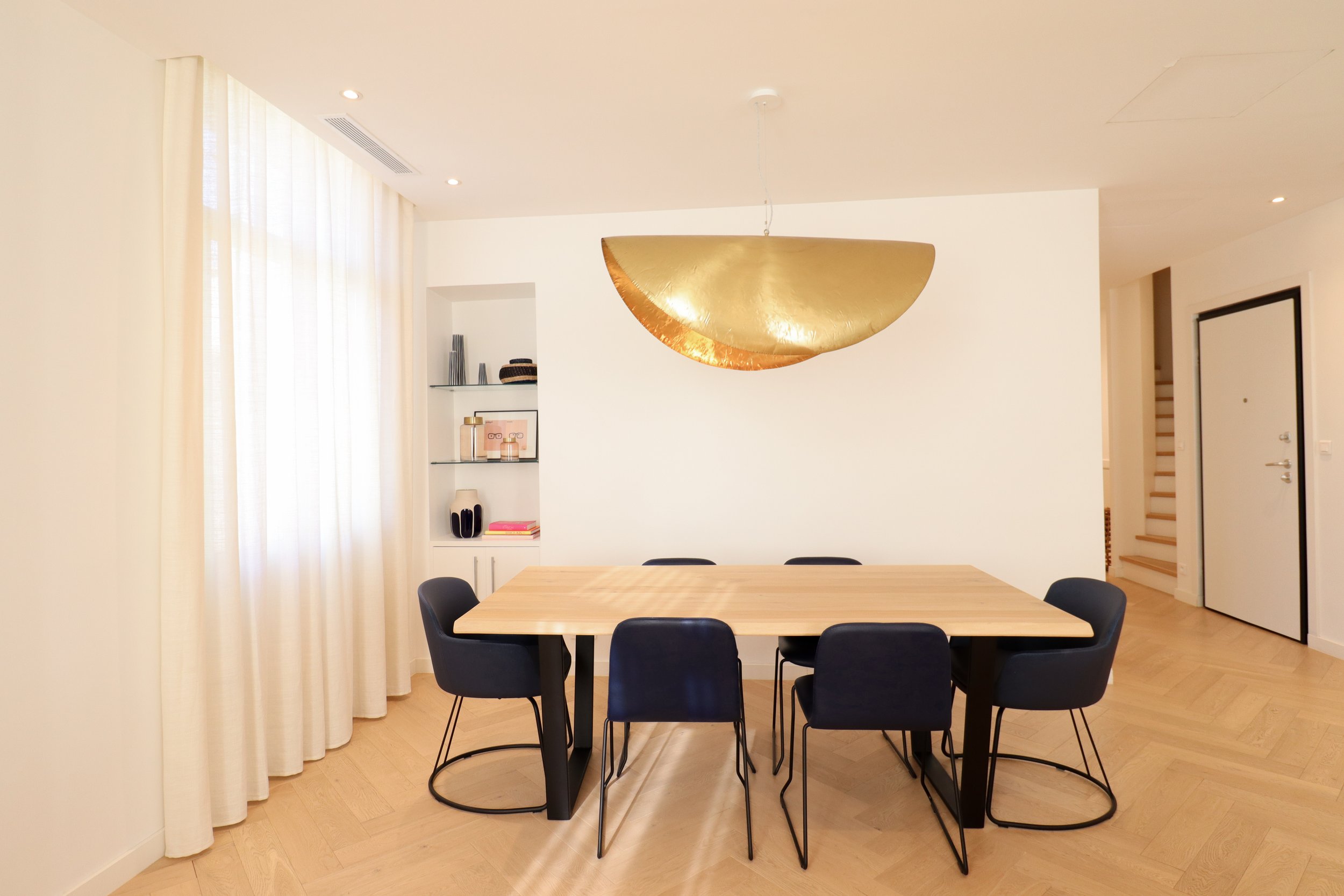 Salle à manger avec une table en bois et six chaises noires. Décor moderne avec une suspension dorée au plafond, étagères murales blanches avec objets décoratifs, et une porte blanche avec cadre noir.