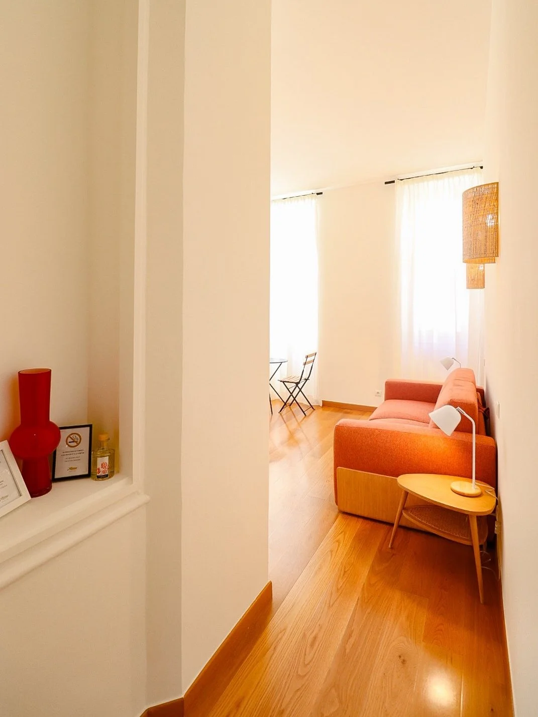 Salon lumineux avec canapé rouge, lampe de chevet, petits meubles en bois, fenêtres avec rideaux blancs, décoration minimaliste.