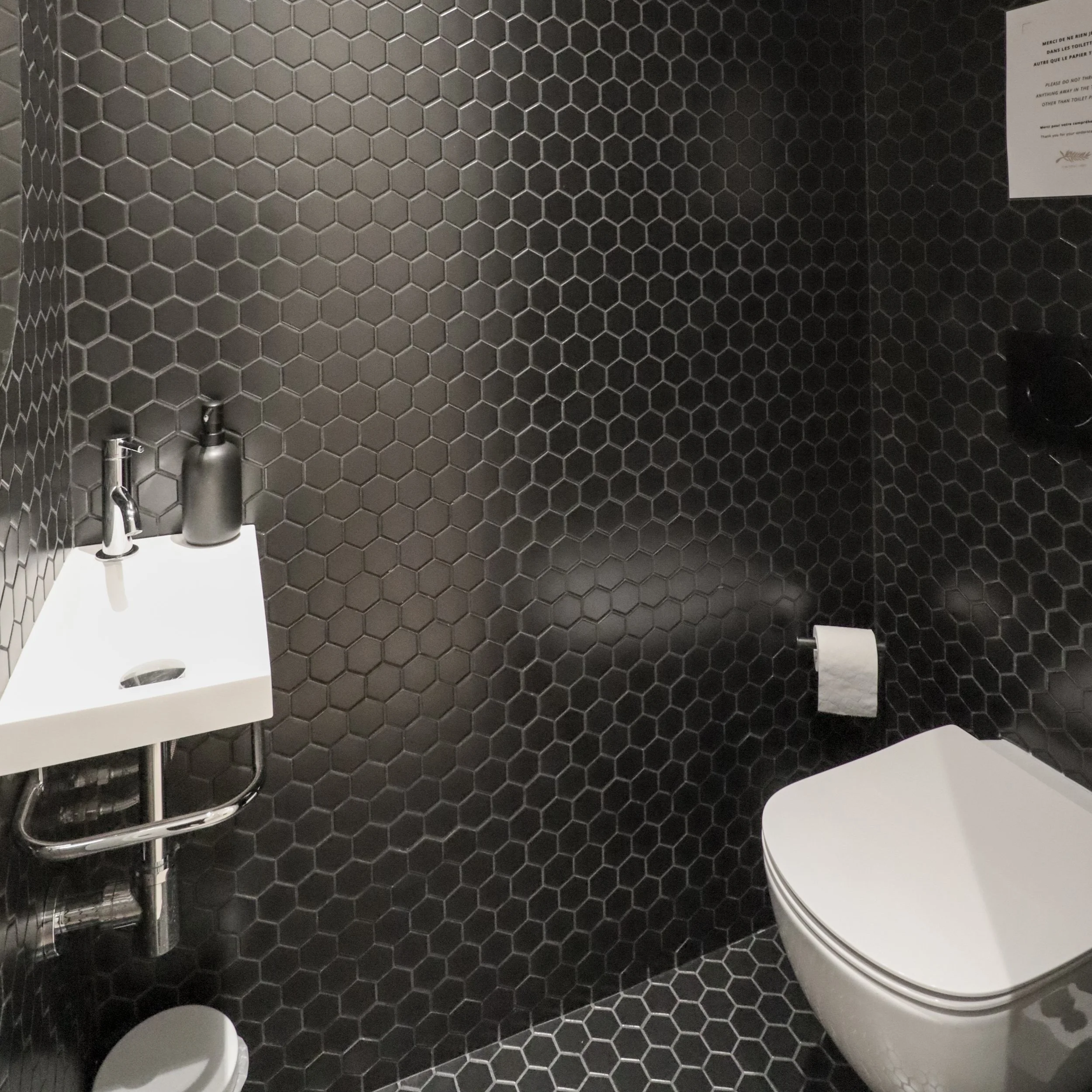 Une cabine de toilettes avec des murs recouverts de carrelage noir en forme de hexagone. Un lavabo blanc avec un distributeur de savon noir et un autre flacon à côté. Un papier toilette est suspendu sur un porte-rouleau. Un papier avec un message est