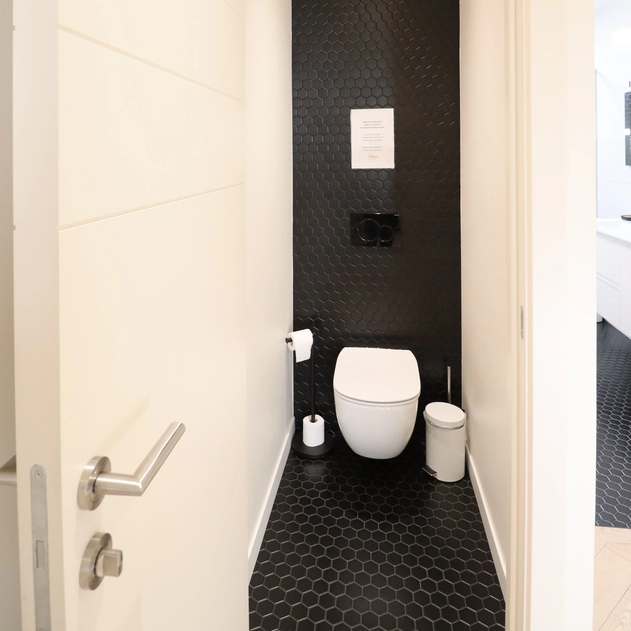 Toilettes dans une salle moderne avec murs en carrelage noir hexagonal, porte partiellement ouverte, et accessoires tels que un porte-papier toilette, un seau, et un panneau d'instructions.