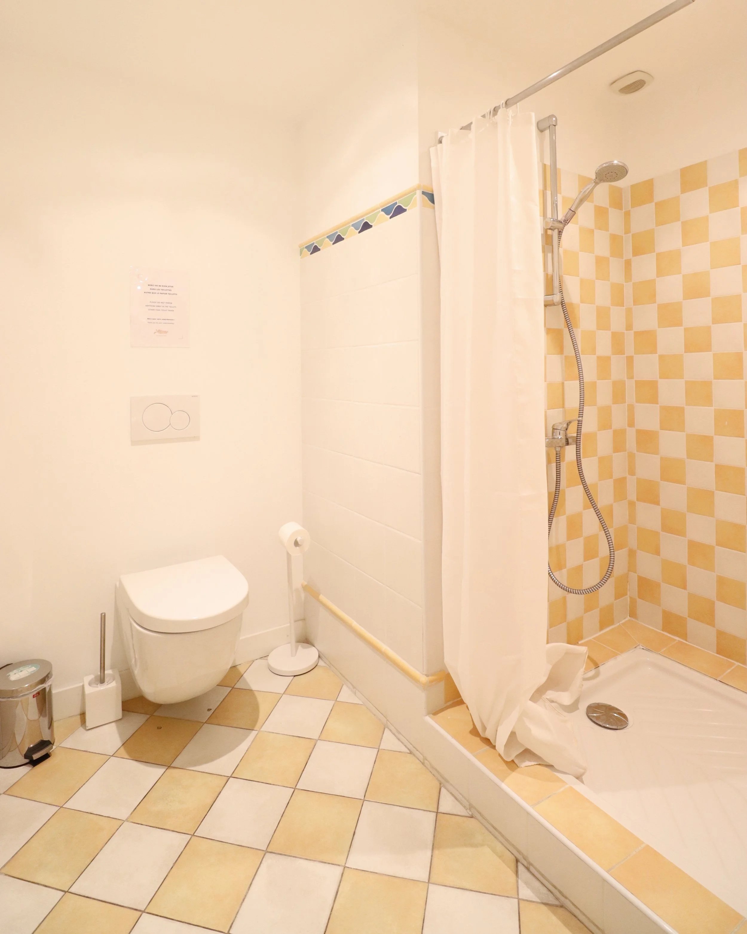 Salle de bain avec toilettes, douche avec rideau blanc, carrelage beige et jaune, accessoires de toilette.