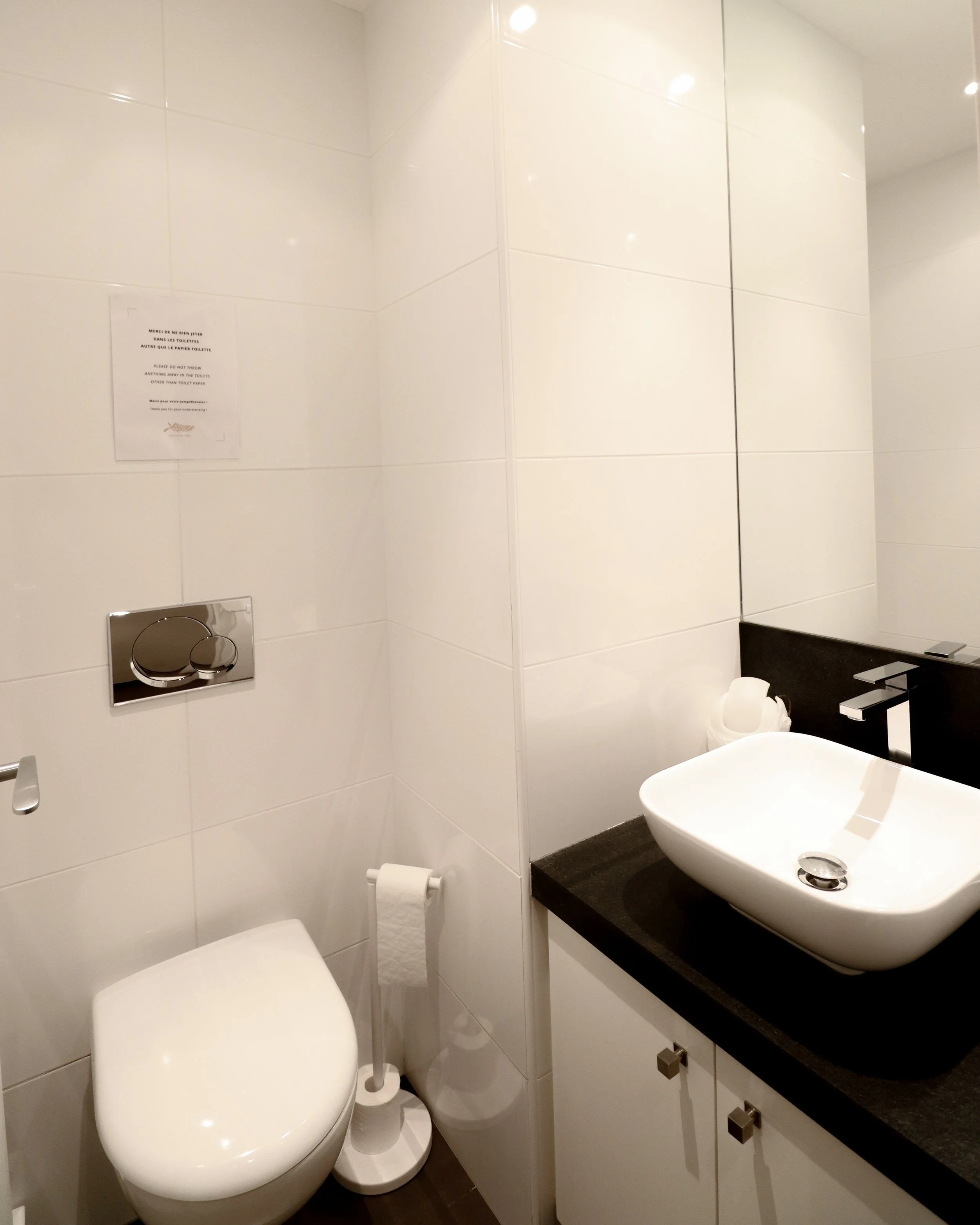 Salle de bain moderne avec un lavabo blanc sur un plan en pierre noire, des toilettes suspendues, des murs en carrelage blanc, un miroir et un robinet noir.