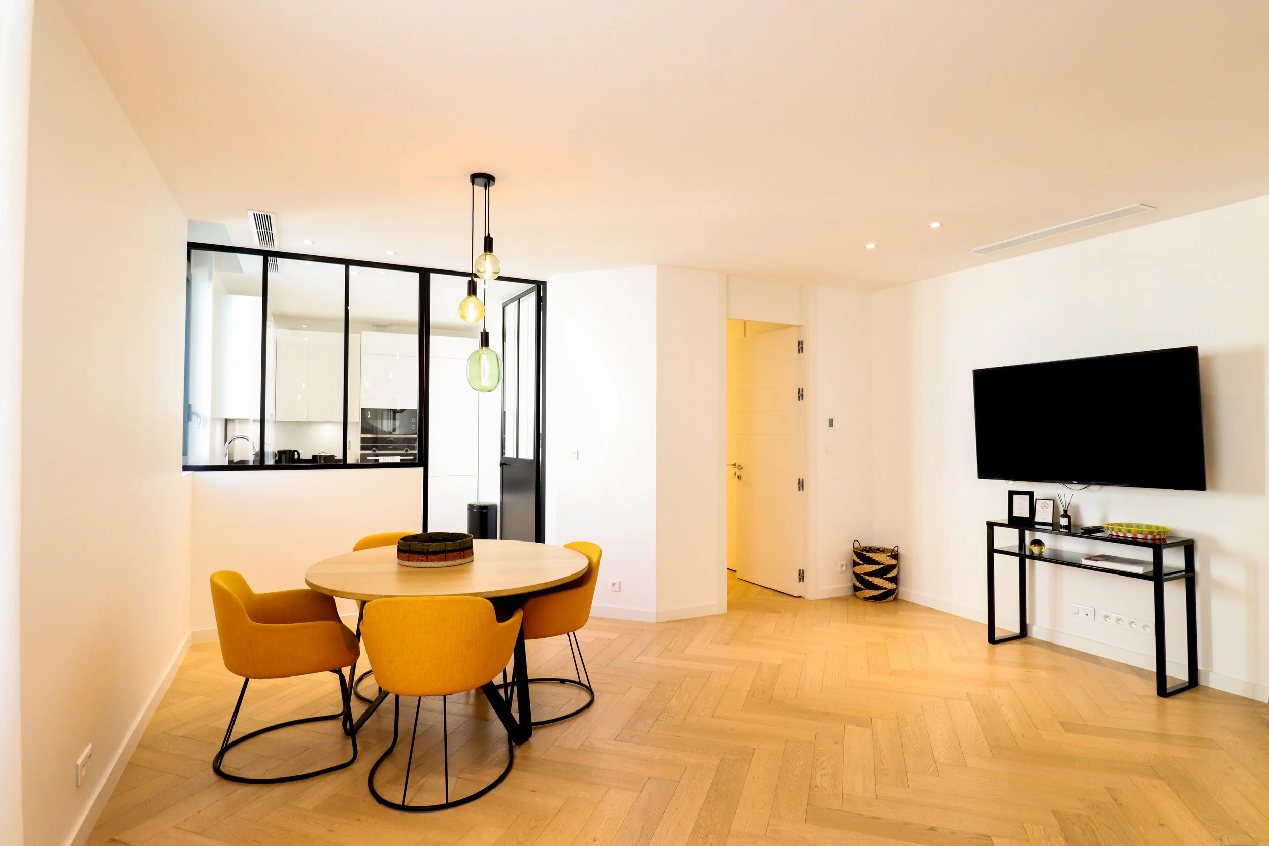 Intérieur d'une salle à manger moderne avec un mur blanc, un plafond beige, un sol en parquet en pente, une table ronde en bois avec des chaises jaunes, un éclairage suspendu avec trois ampoules, une porte donnant sur une cuisine équipée et une télév