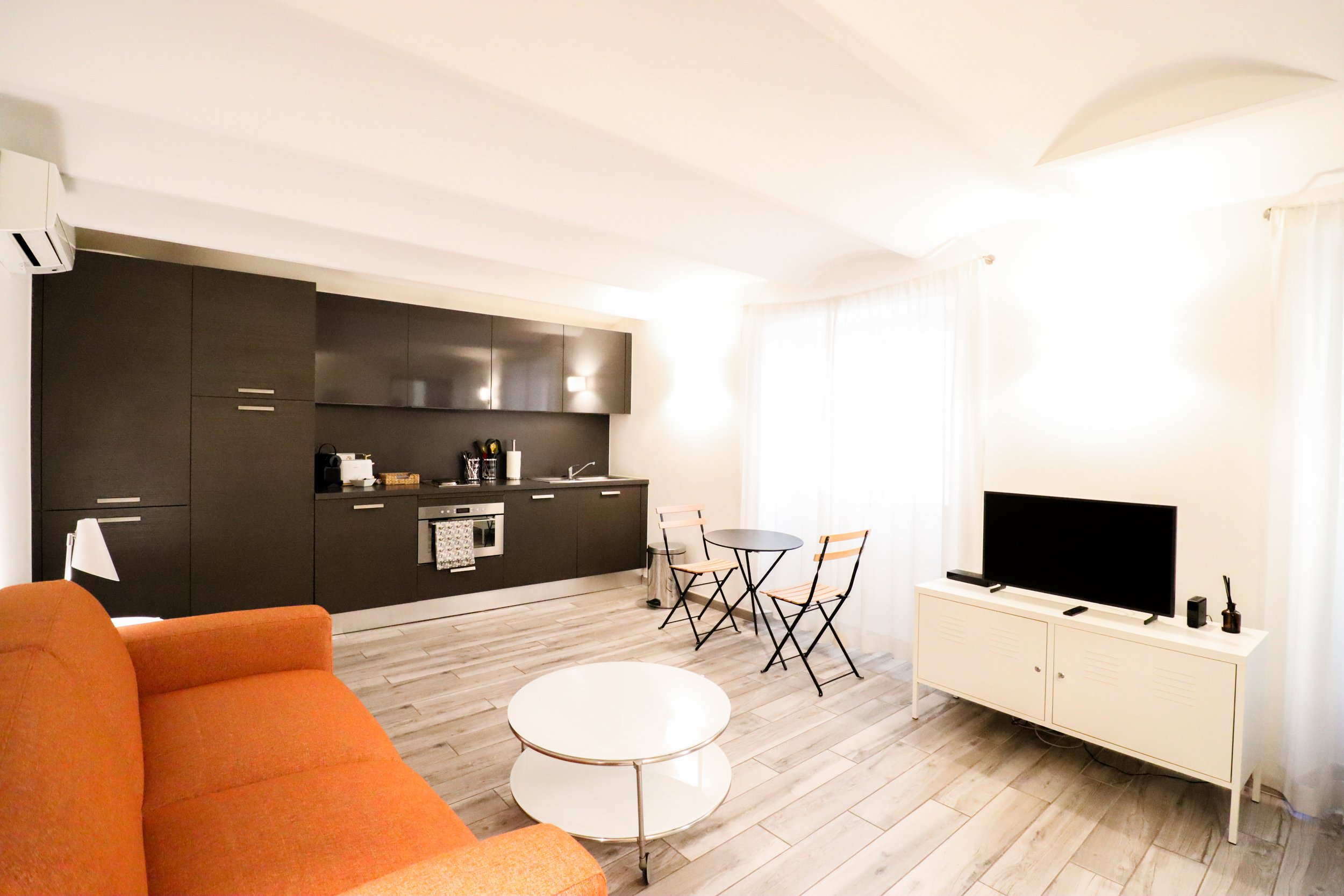 Intérieur de salon avec canapé orange, table ronde blanche, cuisine noire avec armoires, télévision sur meuble blanc et chaise pliante près de fenêtres avec rideaux blancs.