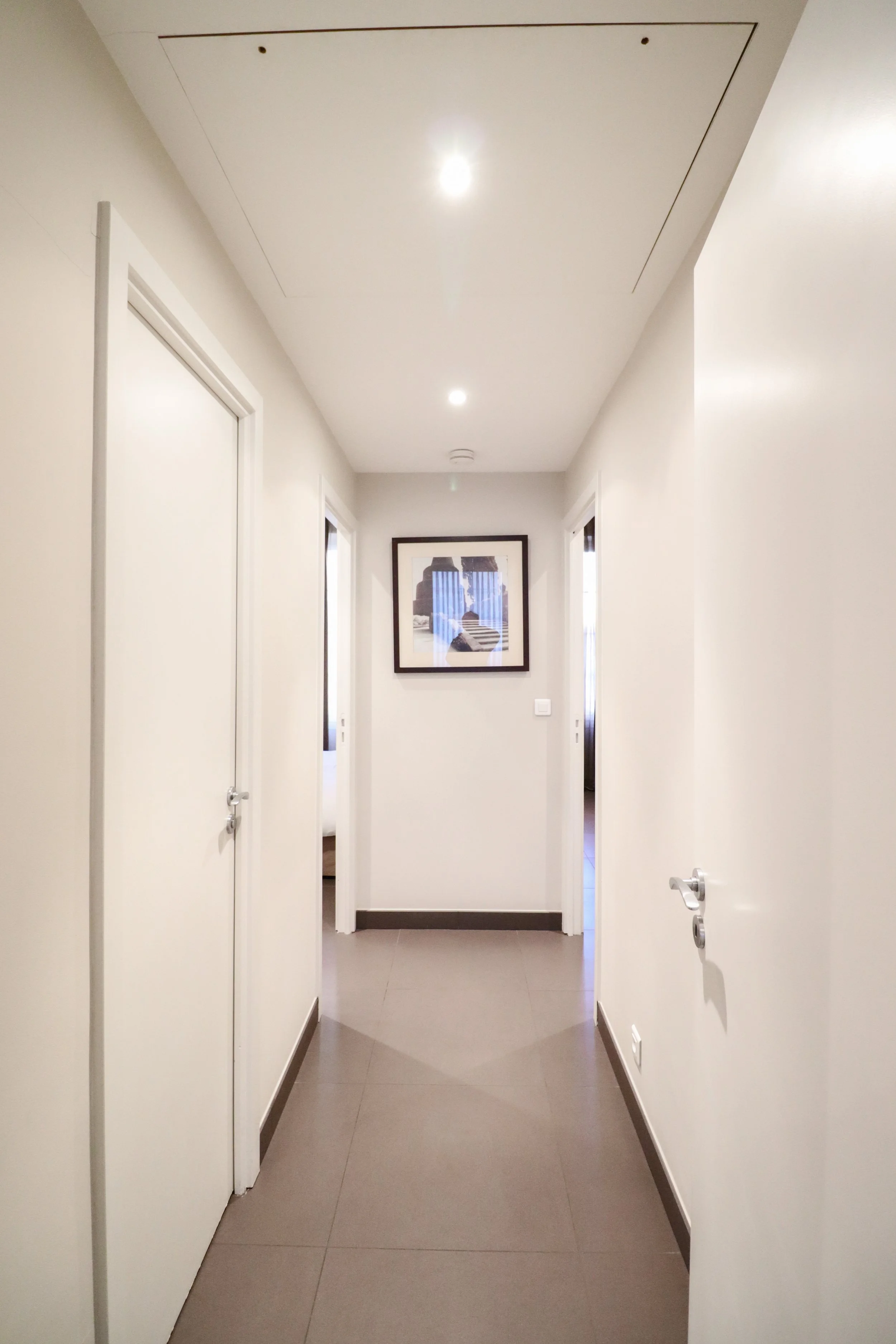 Couloir dans un bâtiment avec des murs blancs, des portes et un tableau suspendu au fond, éclairé par des plafonniers.