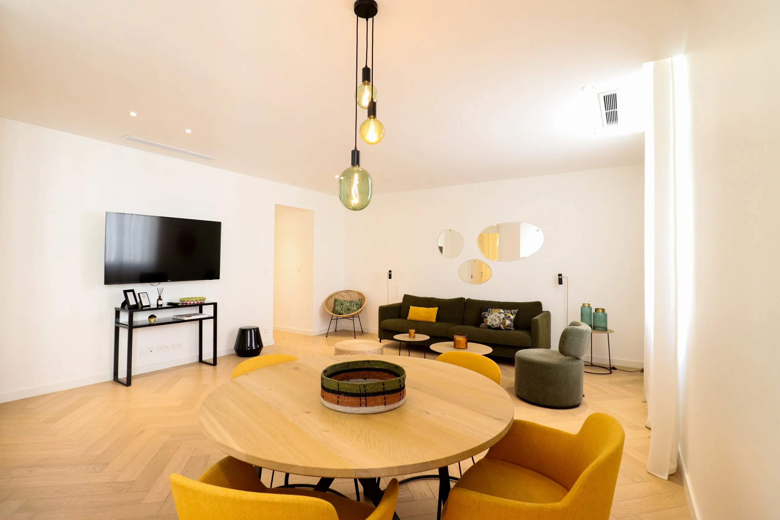Salon moderne avec canapé vert, table ronde en bois avec chaises jaunes, décoration épurée, lampes suspendues en verre, télévision à écran plat, miroirs ovales sur le mur, accessoires décoratifs