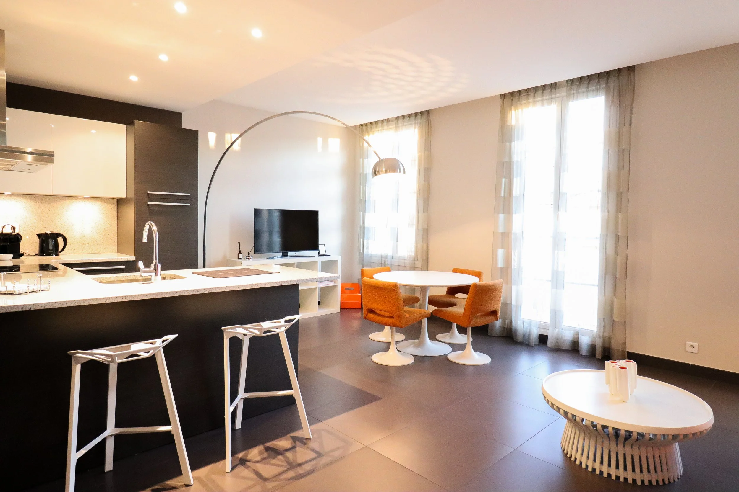 Cuisine moderne avec coin repas, salon et grands fenêtres avec rideaux, mobilier orange, table blanche, plan de travail avec appareils électroménagers, télévision
