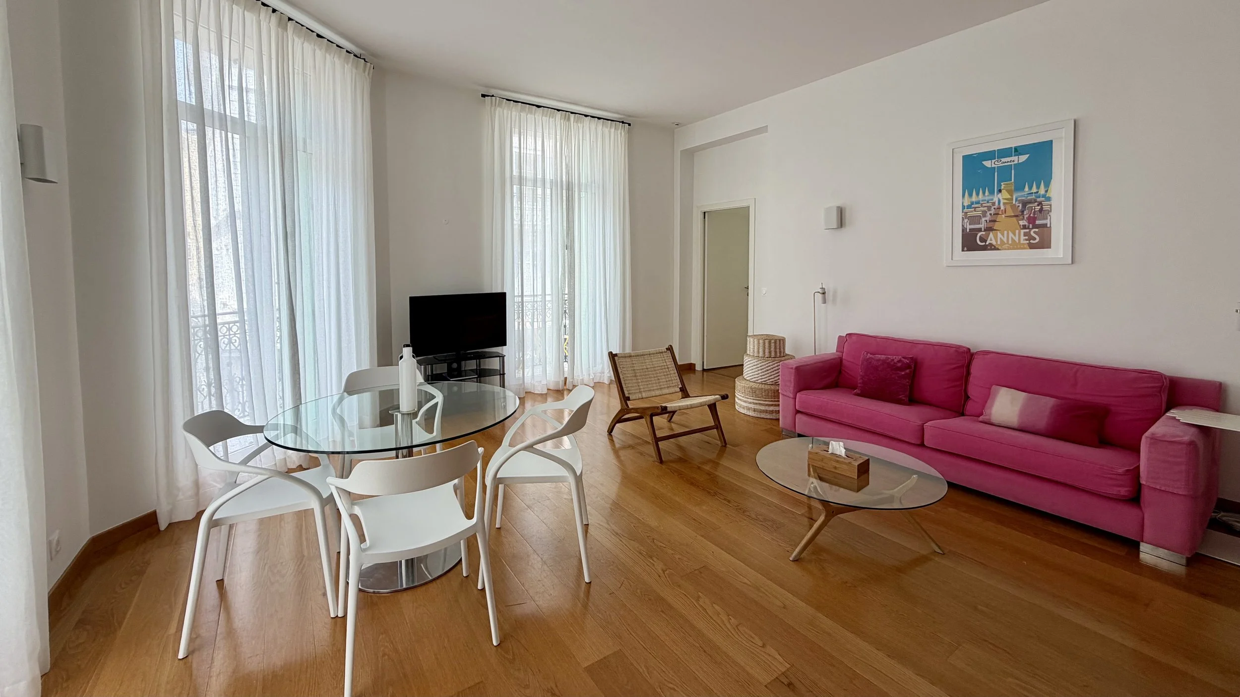 Salon lumineux avec canapé rose, tableau de Cannes, table basse en verre, fauteuil en bois et céramique, télévision, grandes fenêtres avec rideaux blancs, sol en bois.