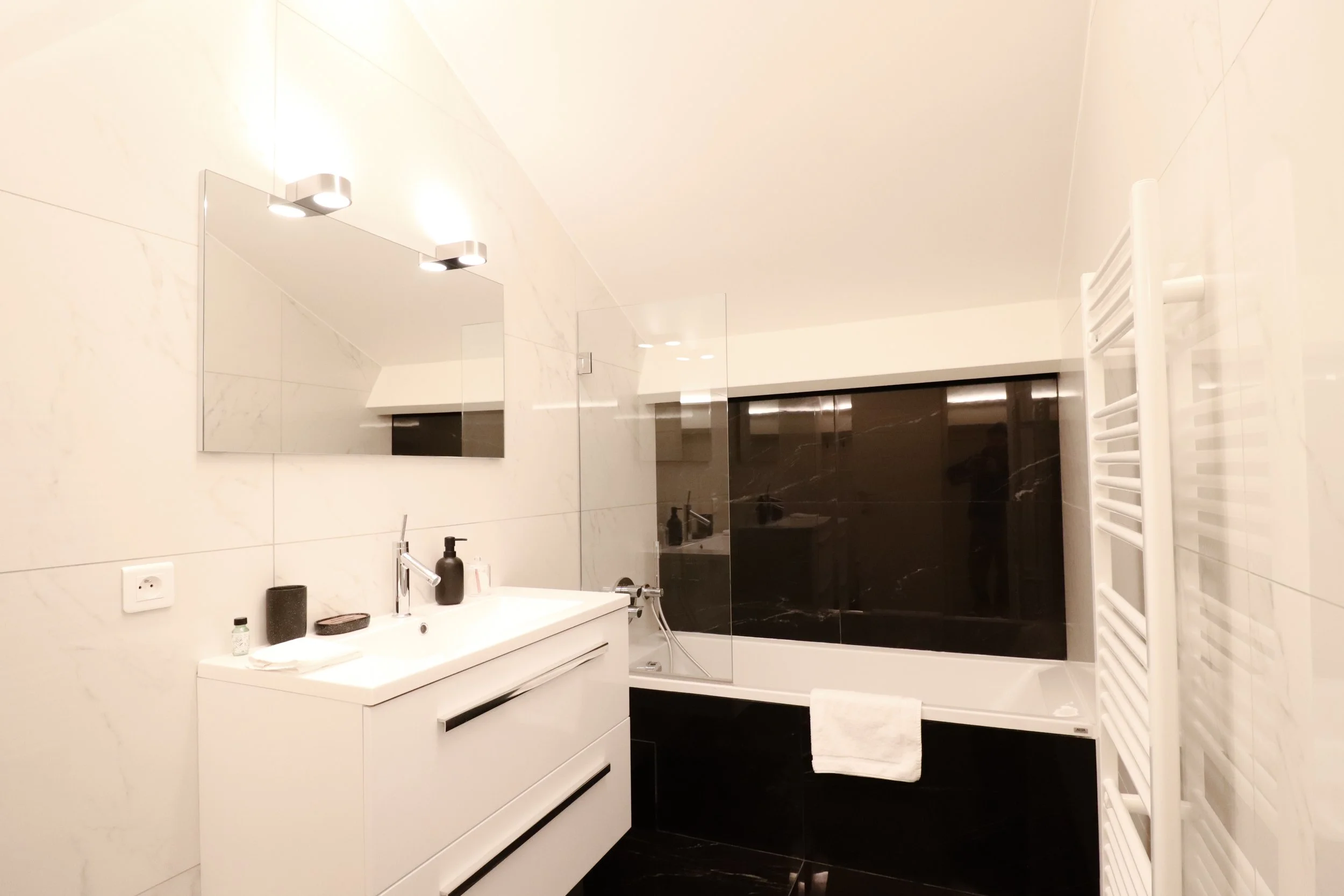 Salle de bain moderne avec murs en marbre blanc, lavabo blanc, miroir rectangulaire, éclairage encastré, radiateur électrique blanc, porte de douche en verre avec cadre noir, et éléments de toilette. Décor minimaliste et lumineux.