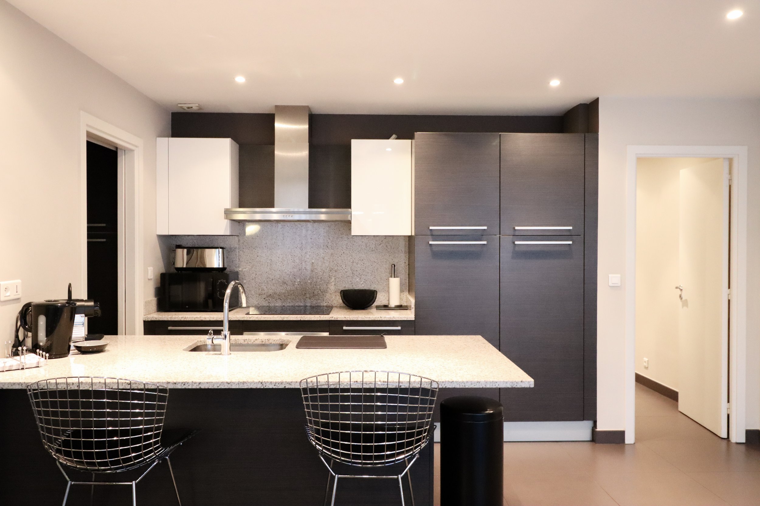 Cuisine moderne avec îlot central, deux chaises en fil noir, placards gris foncé et blancs, hotte en acier inoxydable, murs gris clair, éclairage encastré au plafond.