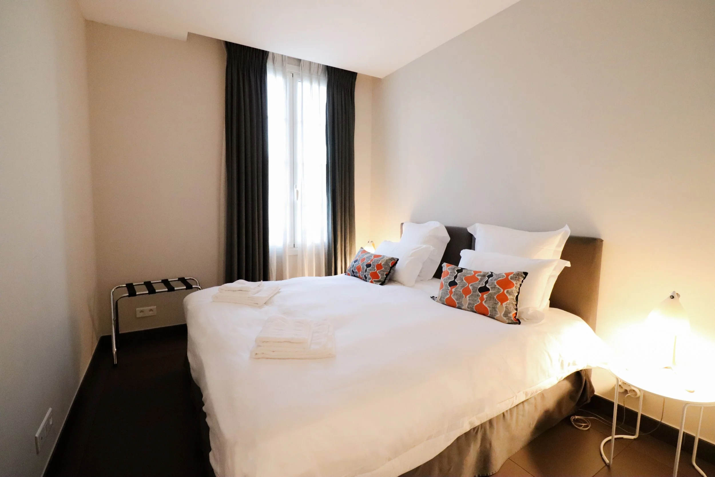 Chambre d'hôtel avec un lit double, oreillers, draps blancs, une fenêtre avec rideaux foncés, lampe de lecture, et une petite table à côté du lit.
