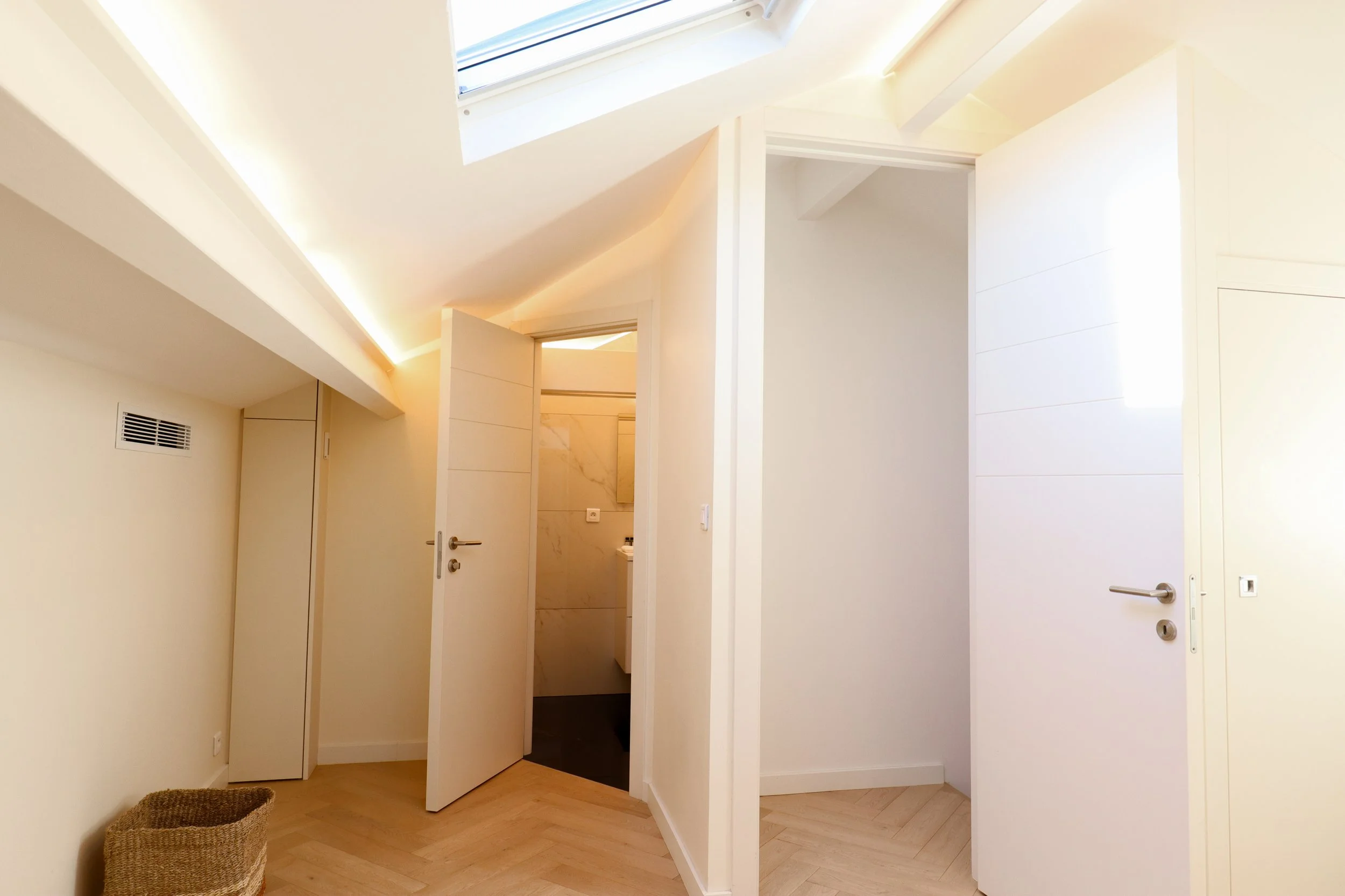 Intérieur d'une pièce avec des murs blancs, un plafond en pente avec une fenêtre de toit, une porte ouverte menant à une salle de bain, un panier en osier dans un coin.