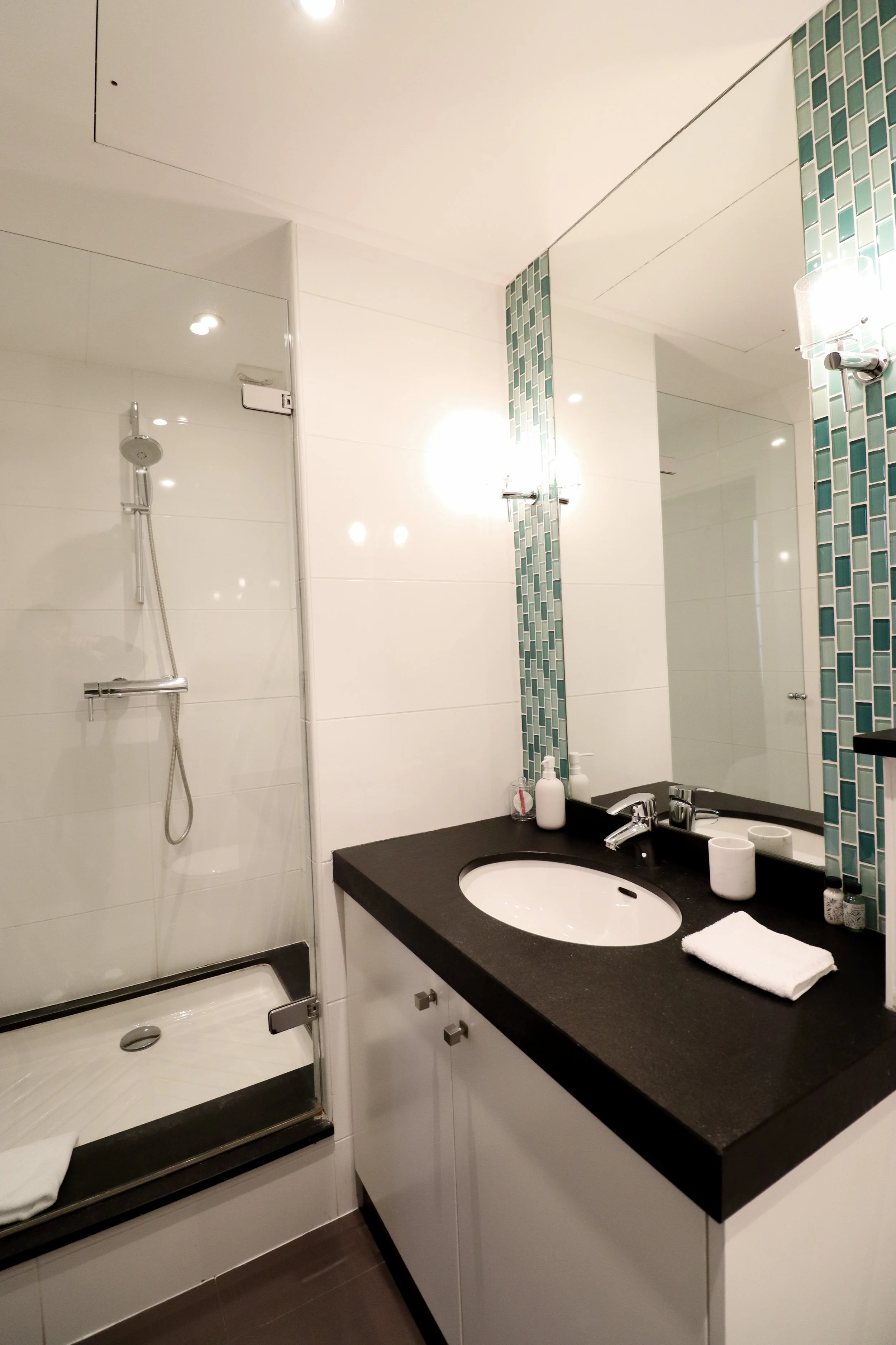 Une salle de bain moderne avec une cabine de douche, un lavabo avec un miroir au-dessus, des accessoires de toilette, une serviette blanche, des murs en carrelage blanc et des détails en mosaïque turquoise.