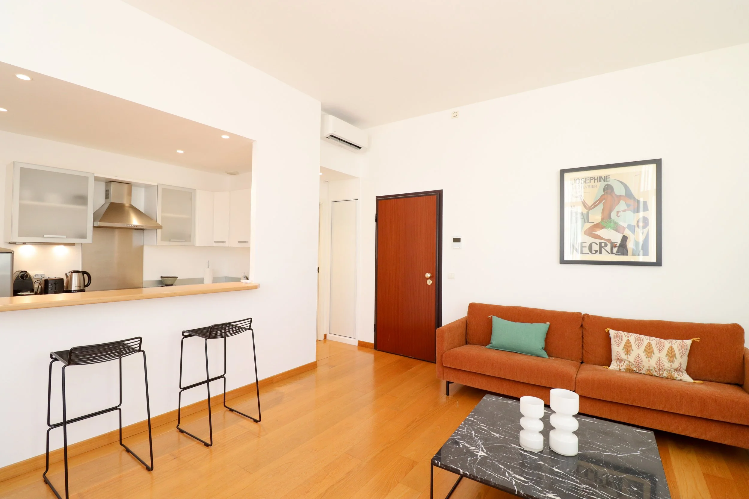 Intérieur moderne d'un salon et cuisine, comprenant un canapé orange avec coussins verts et imprimés, une table basse en marbre noir avec bougies blanches, un mur blanc avec une affiche encadrée et une porte en bois.