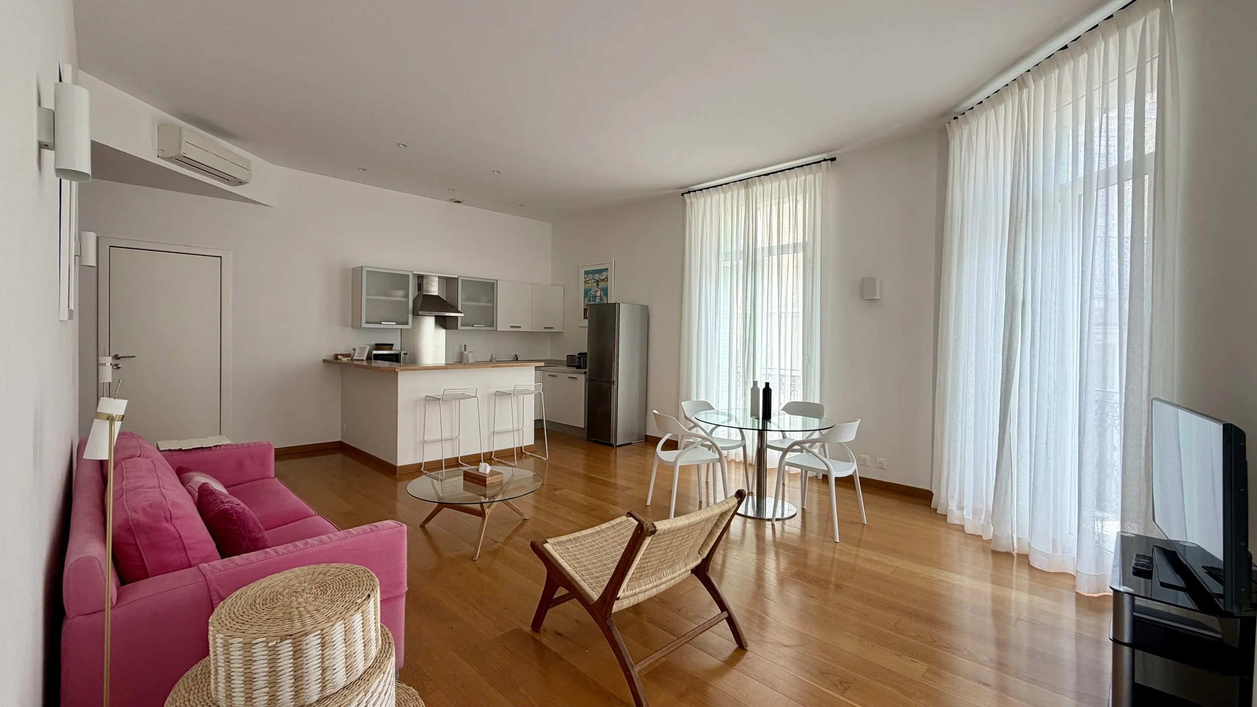 Intérieur d'un appartement avec un salon et une cuisine ouverts, deux grandes fenêtres avec rideaux blancs, un canapé rose, une table basse en verre, un fauteuil en bois avec coussin, un réfrigérateur, une table ronde avec quatre chaises, une télévision sur un meuble, et un mur décoré d'une photo ou peinture.
