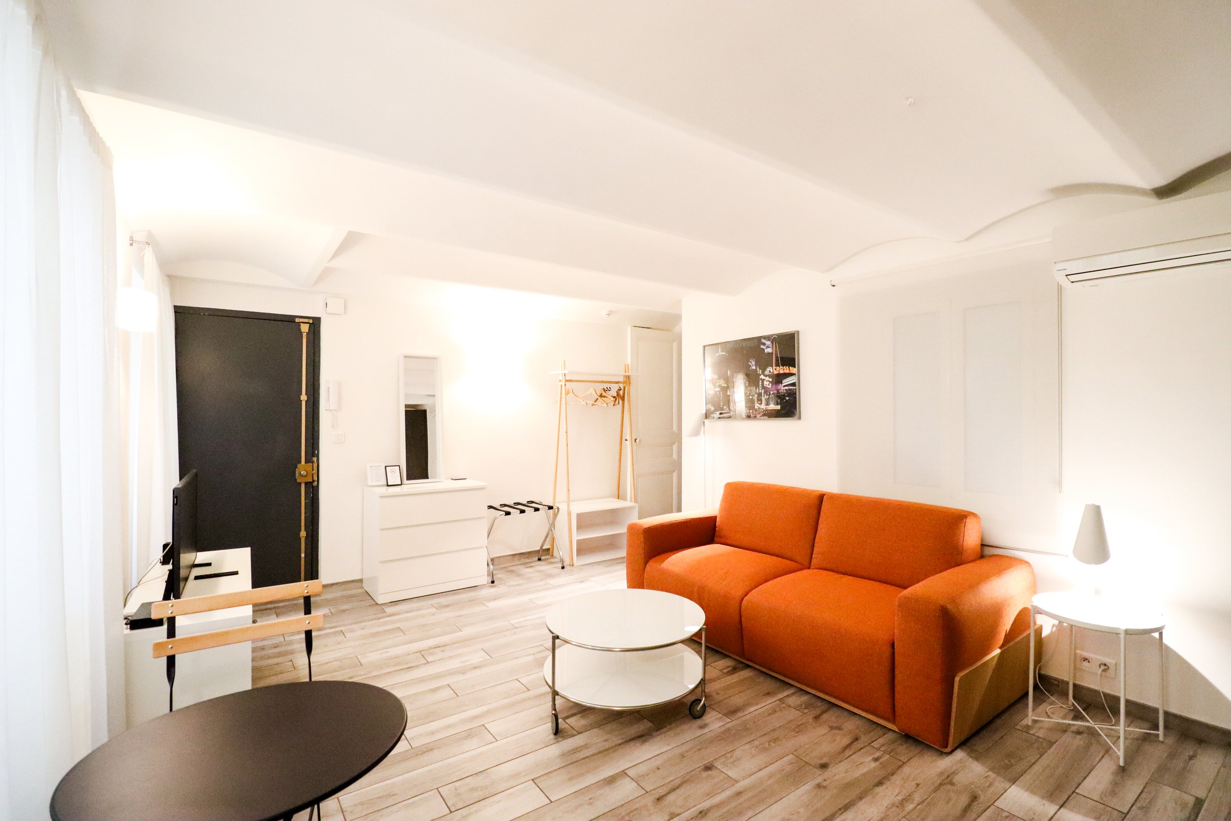 Un salon lumineux avec un canapé orange, une table basse ronde en verre, un mur blanc avec un tableau, un porte-manteau en bois, une télévision, un radiateur et un ventilateur de plafond.